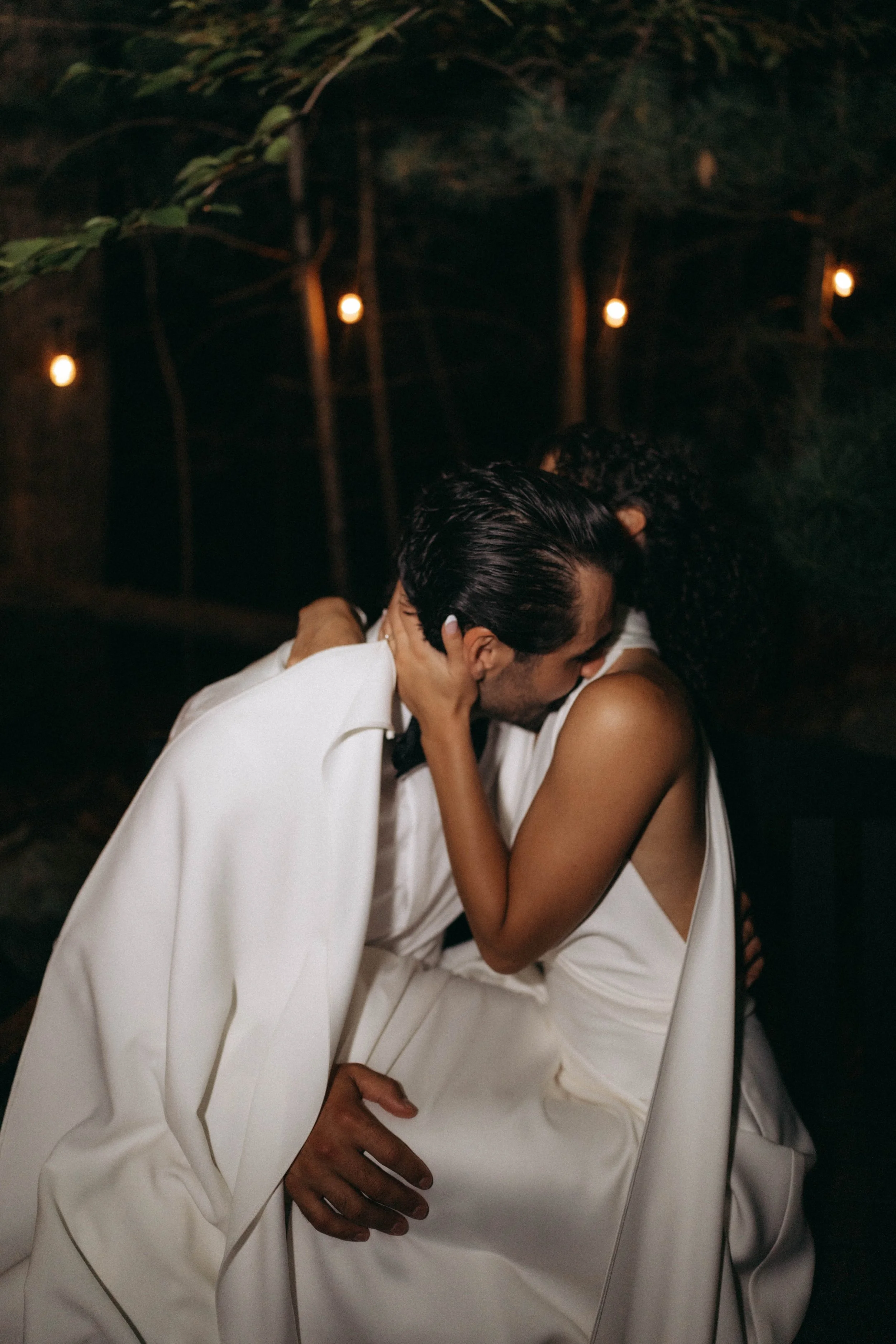 A couple wearing white formal attire, embracing each other, in a dark outdoor setting with string lights overhead.