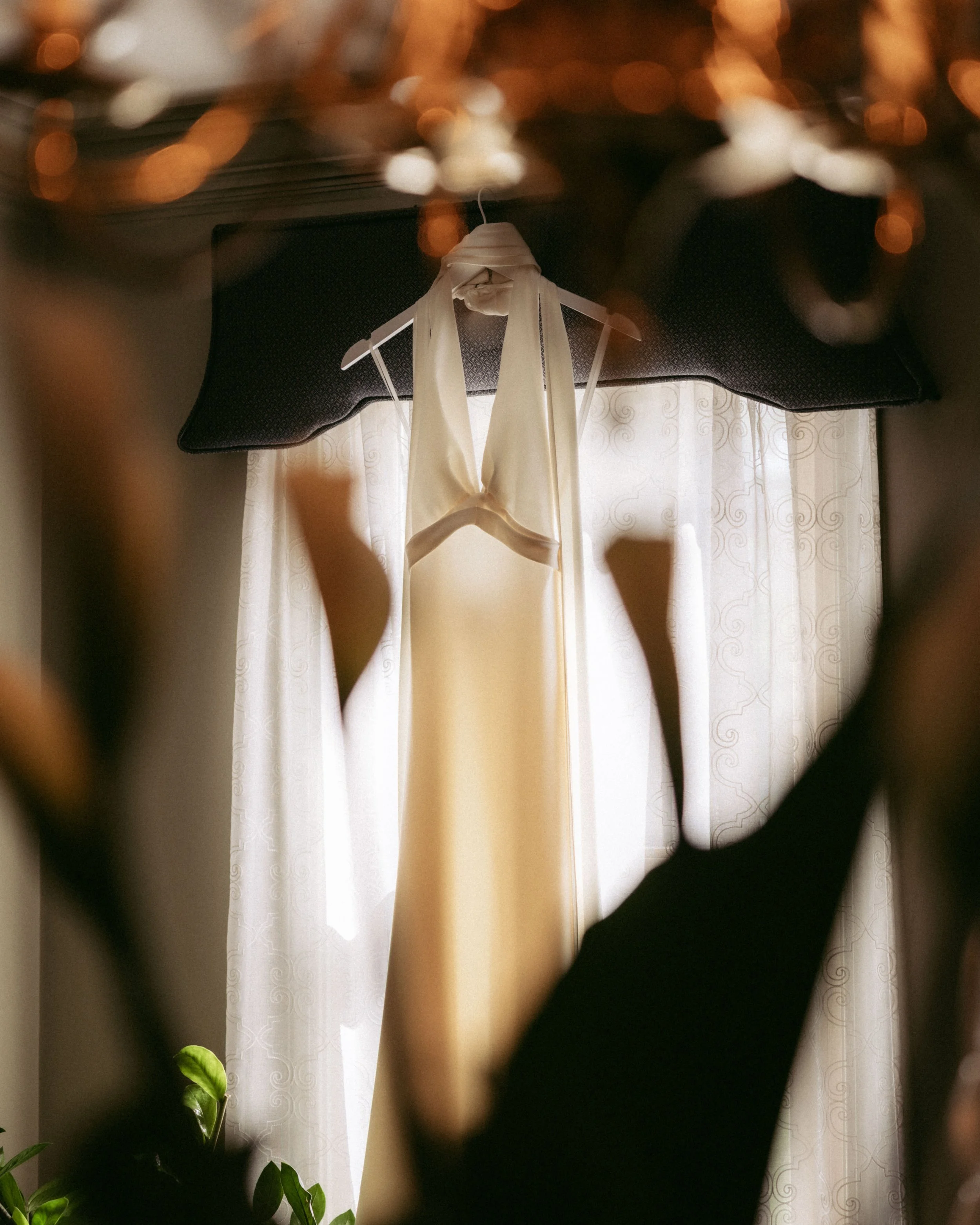 A dress hanging on a hanger in front of a window with curtains, with blurred dark and light foliage in the foreground.