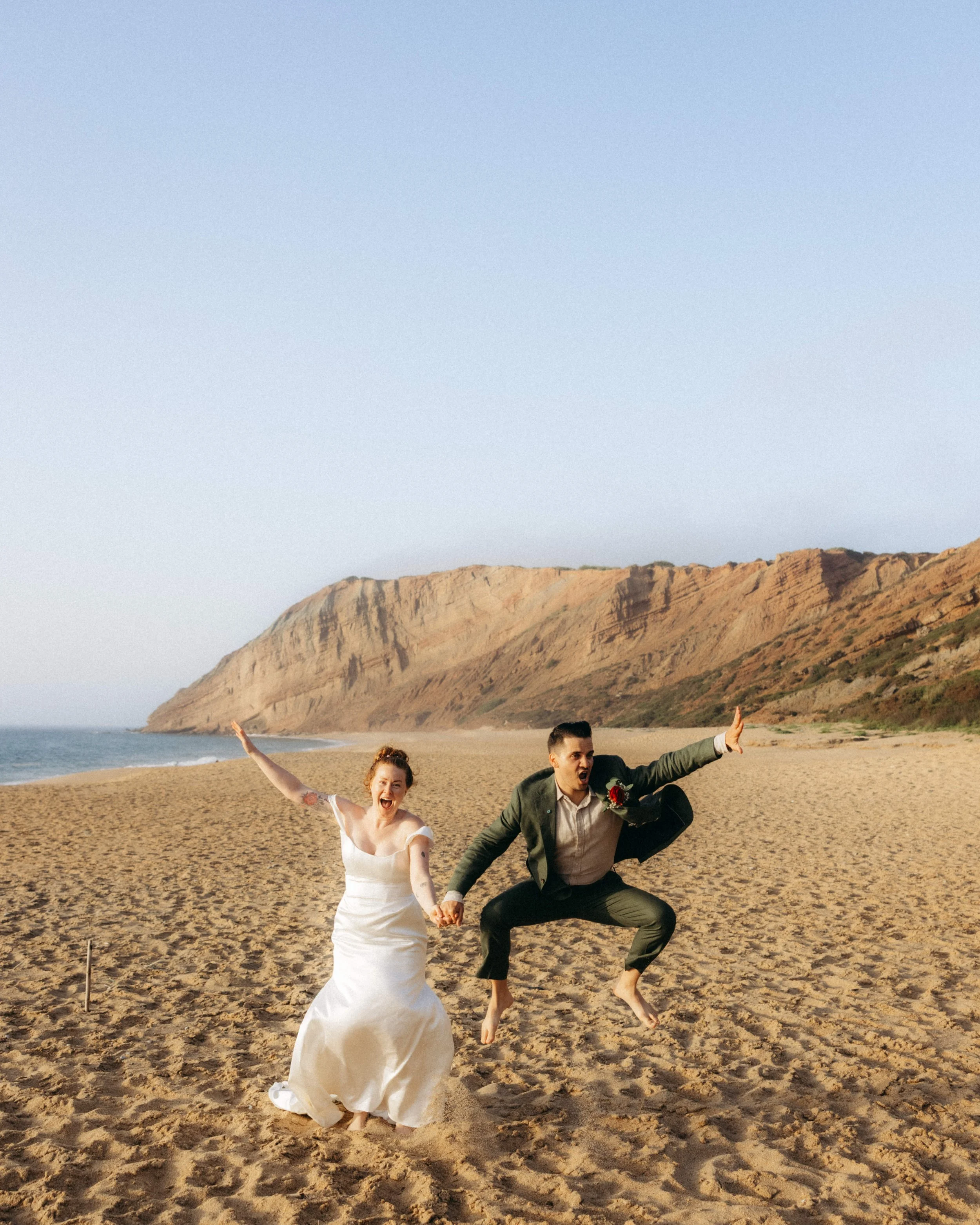 A woman in a white wedding dress and a man in a green suit are on a sandy beach, holding hands and playfully jumping or running. They appear joyful and energetic, with a backdrop of cliffs and the ocean.