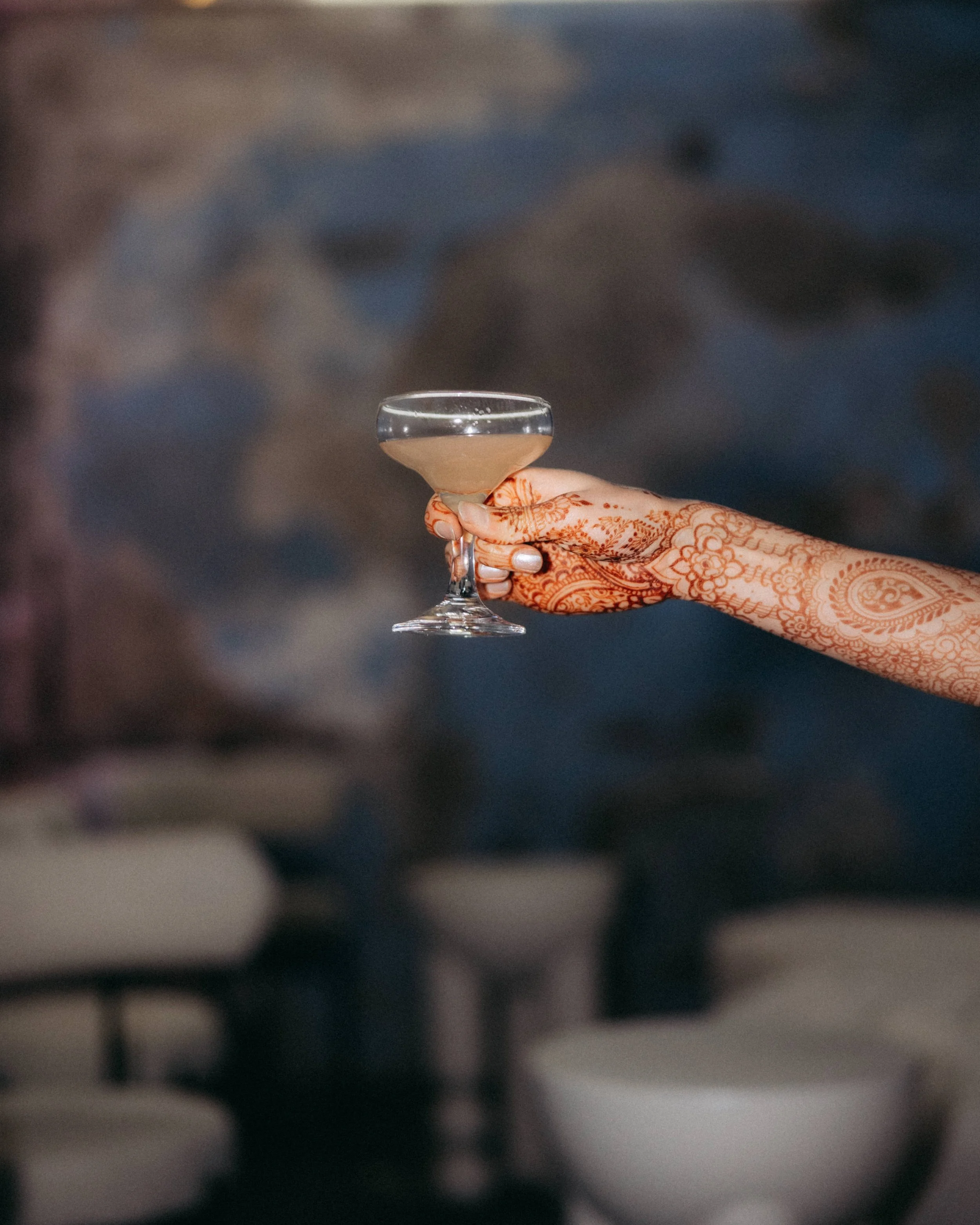 A hand with henna designs holding a clear glass of champagne against a blurred background.