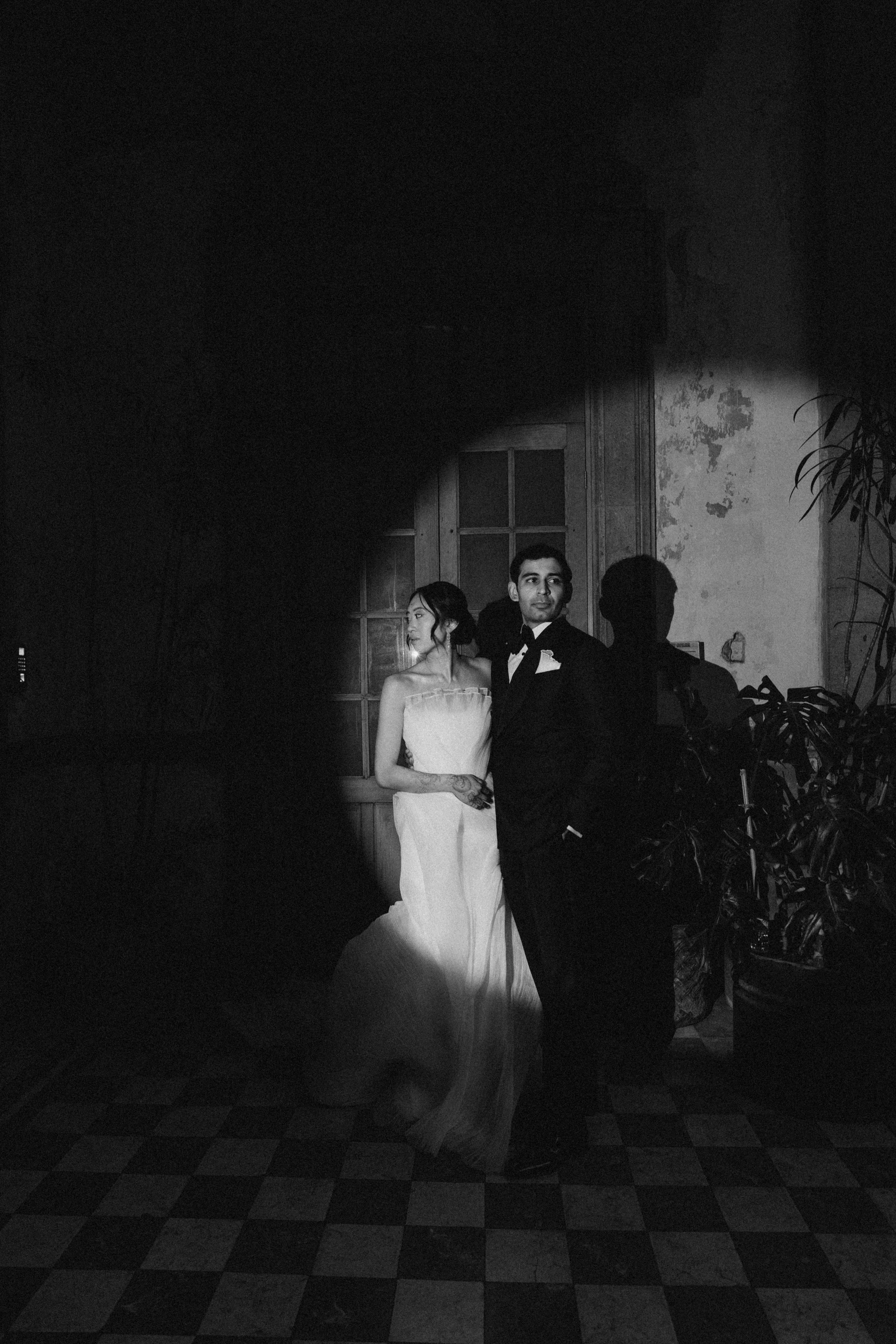 A bride and groom dancing at their wedding reception in a dimly lit room with a checkered floor, a wall with peeling paint, and some plants.