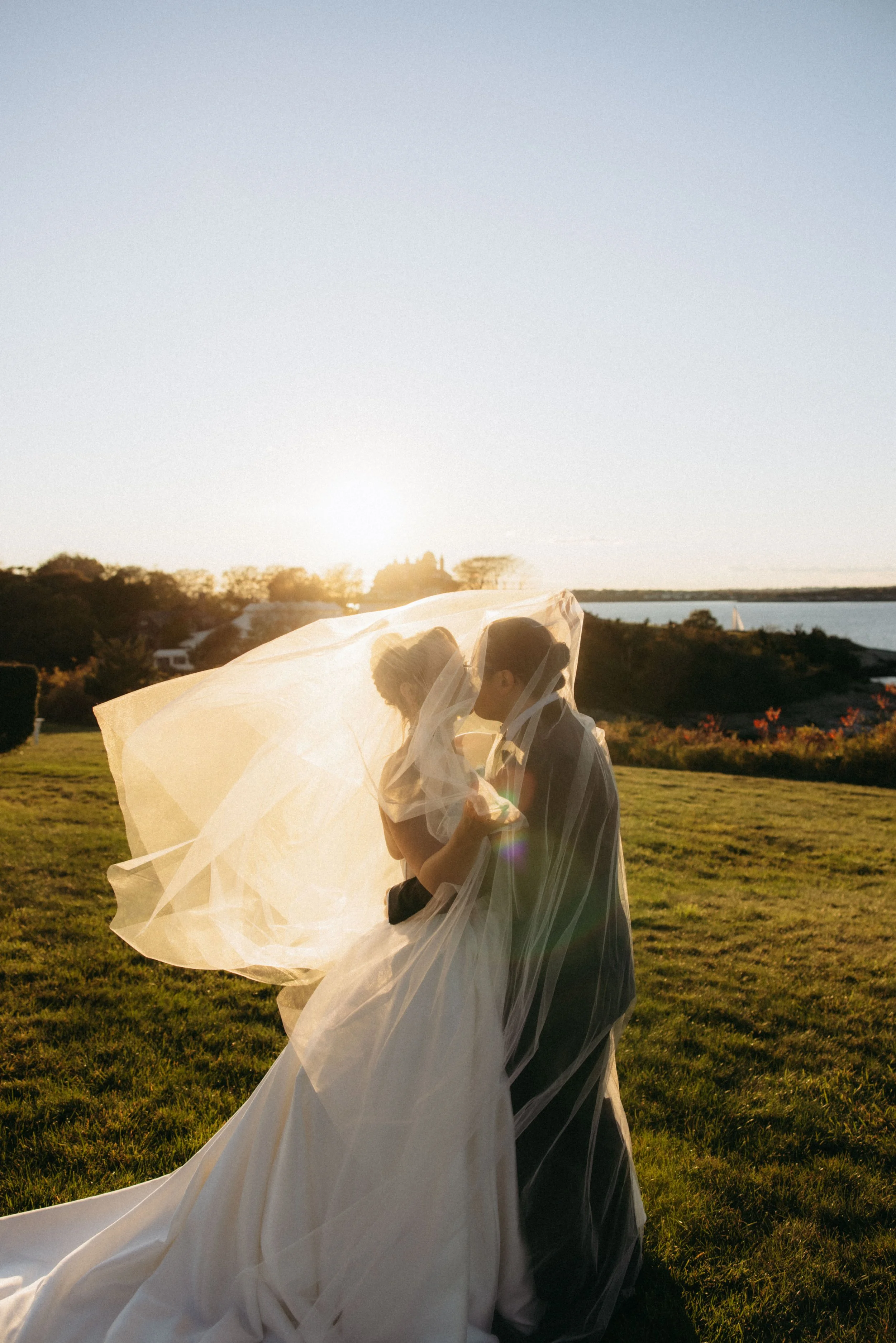 A couple dressed in wedding attire embracing outdoors during sunset, with the bride holding a veil that flows around them, lush green grass, trees, a body of water, and a sailboat in the distance.