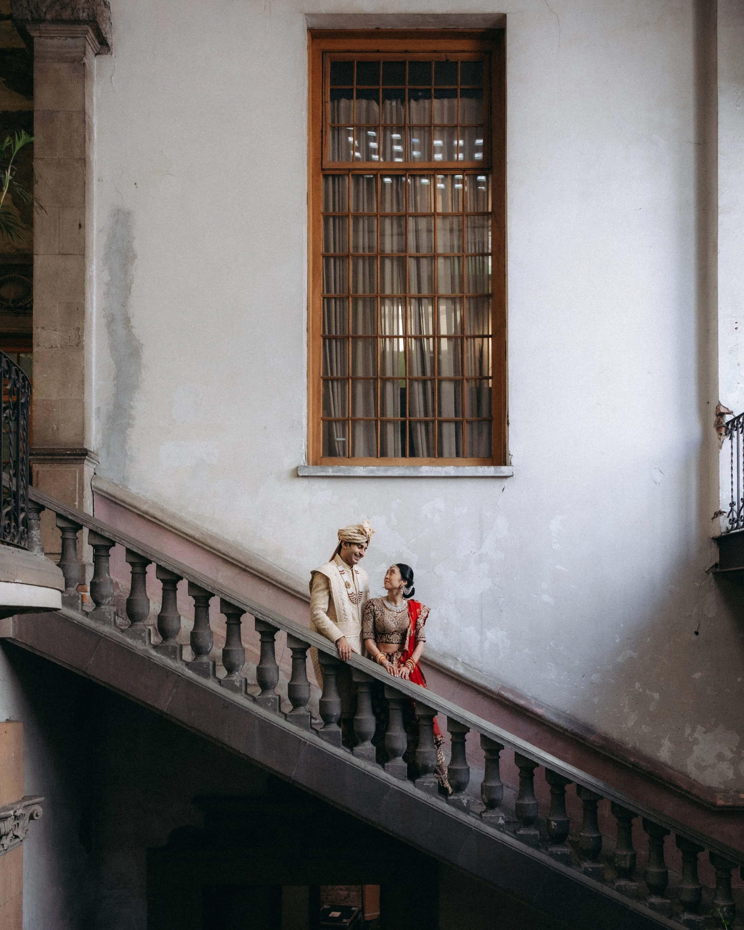 A man and woman in traditional Indian attire standing on a staircase inside a building, with a large window and white wall behind them.
