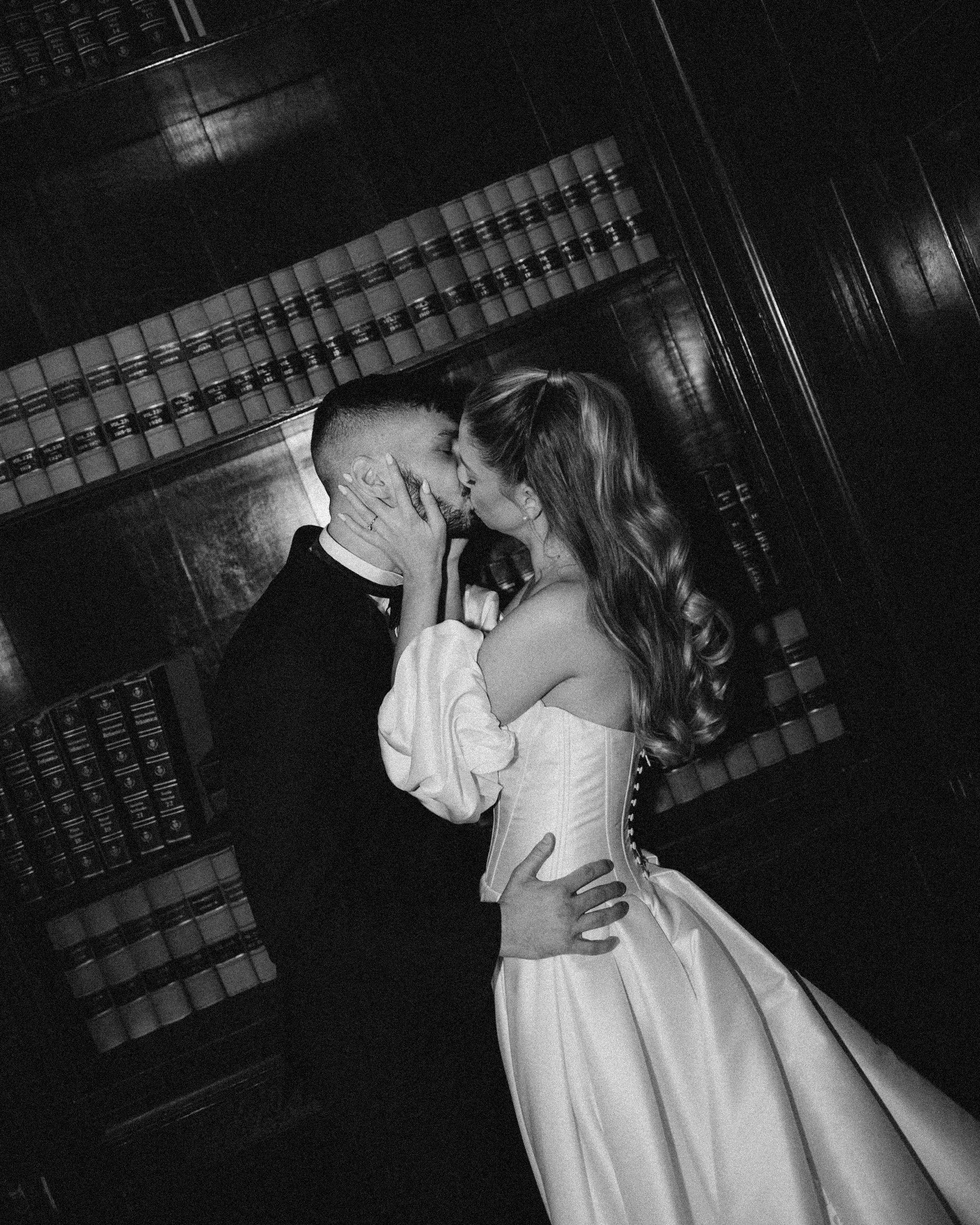 A couple dressed in formal attire sharing an intimate kiss in a dimly lit room with wooden furniture and a shelf of books in the background.