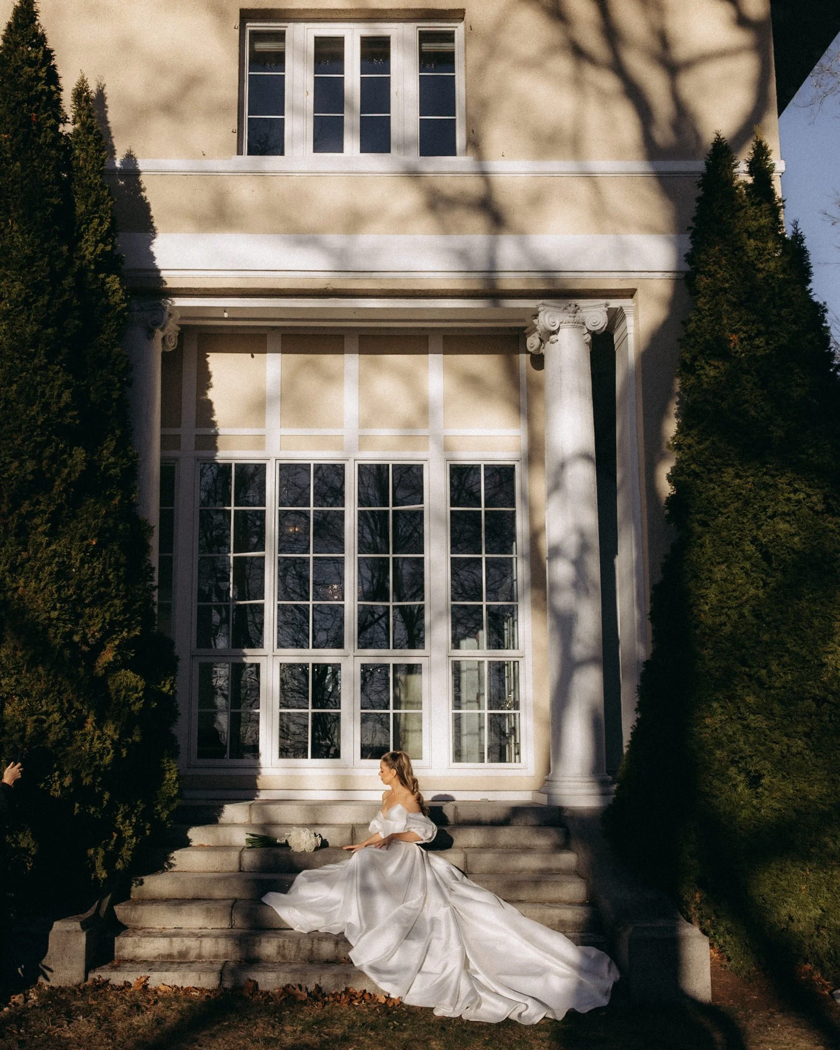 A bride in a white wedding dress sitting on steps outside a large house with tall windows and trees casting shadows.