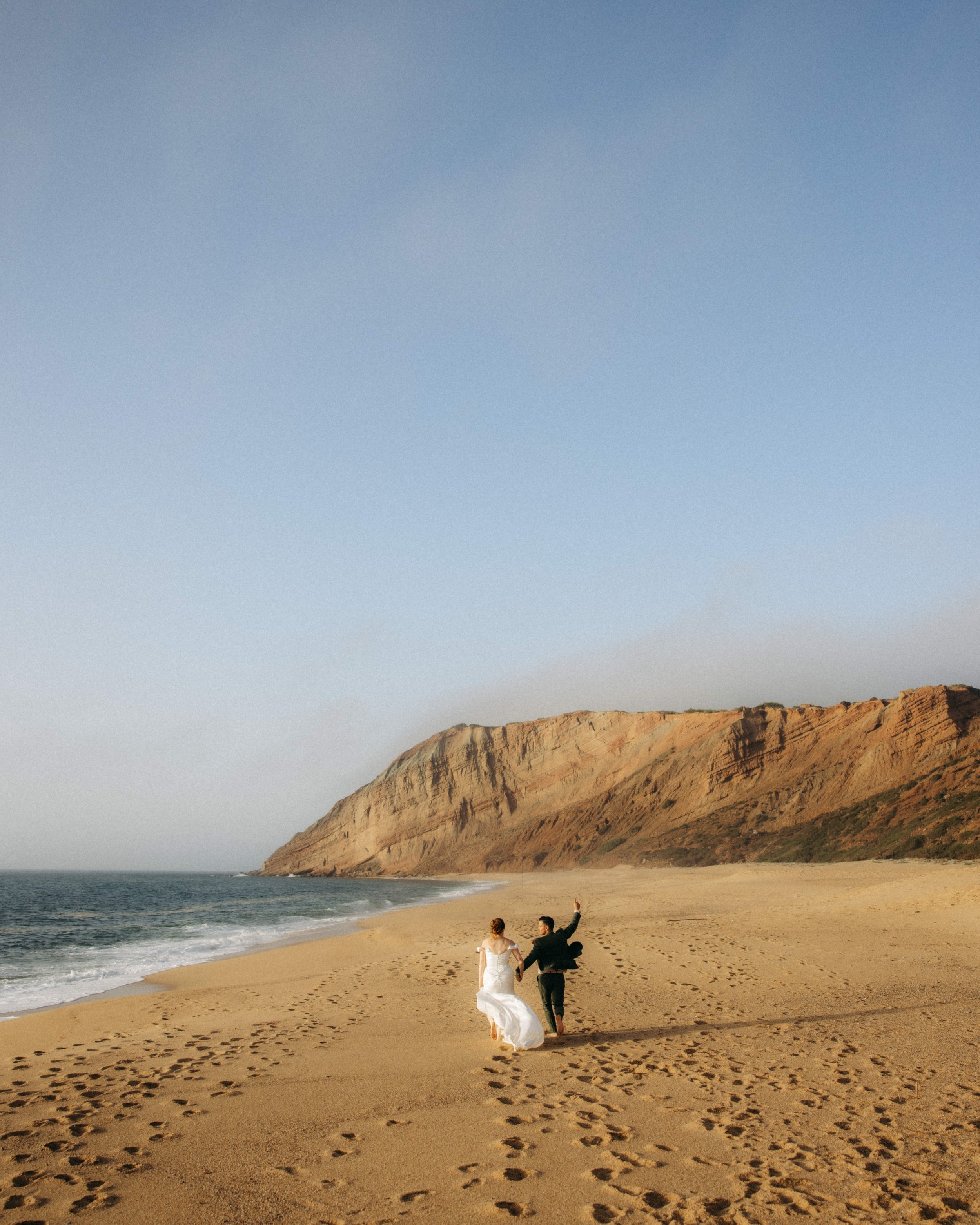 A newlywed couple walking on the beach at sunset, with cliffs in the background and footprints in the sand.