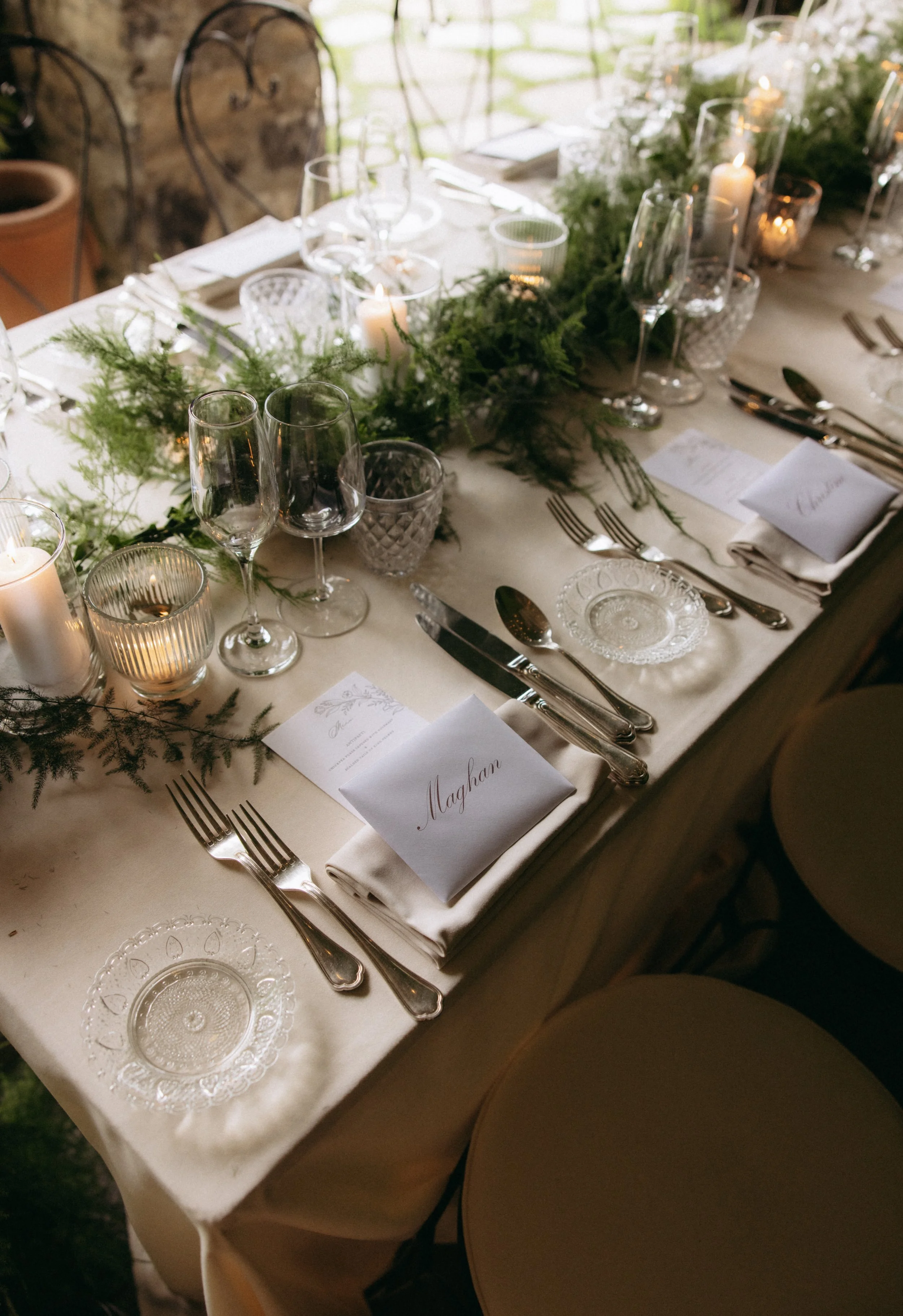 A formal dining table set with glassware, cutlery, white napkins, and small candles, decorated with greenery and a place card that reads "Maghan."