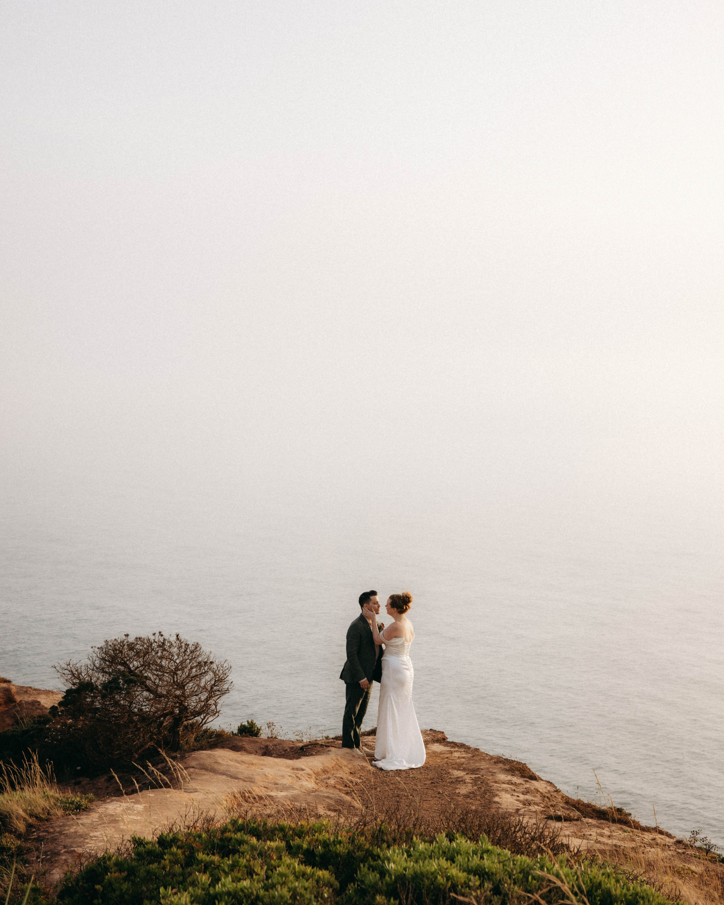 A couple in wedding attire standing on a cliff overlooking the ocean, with sparse vegetation and a cloudy sky in the background.
