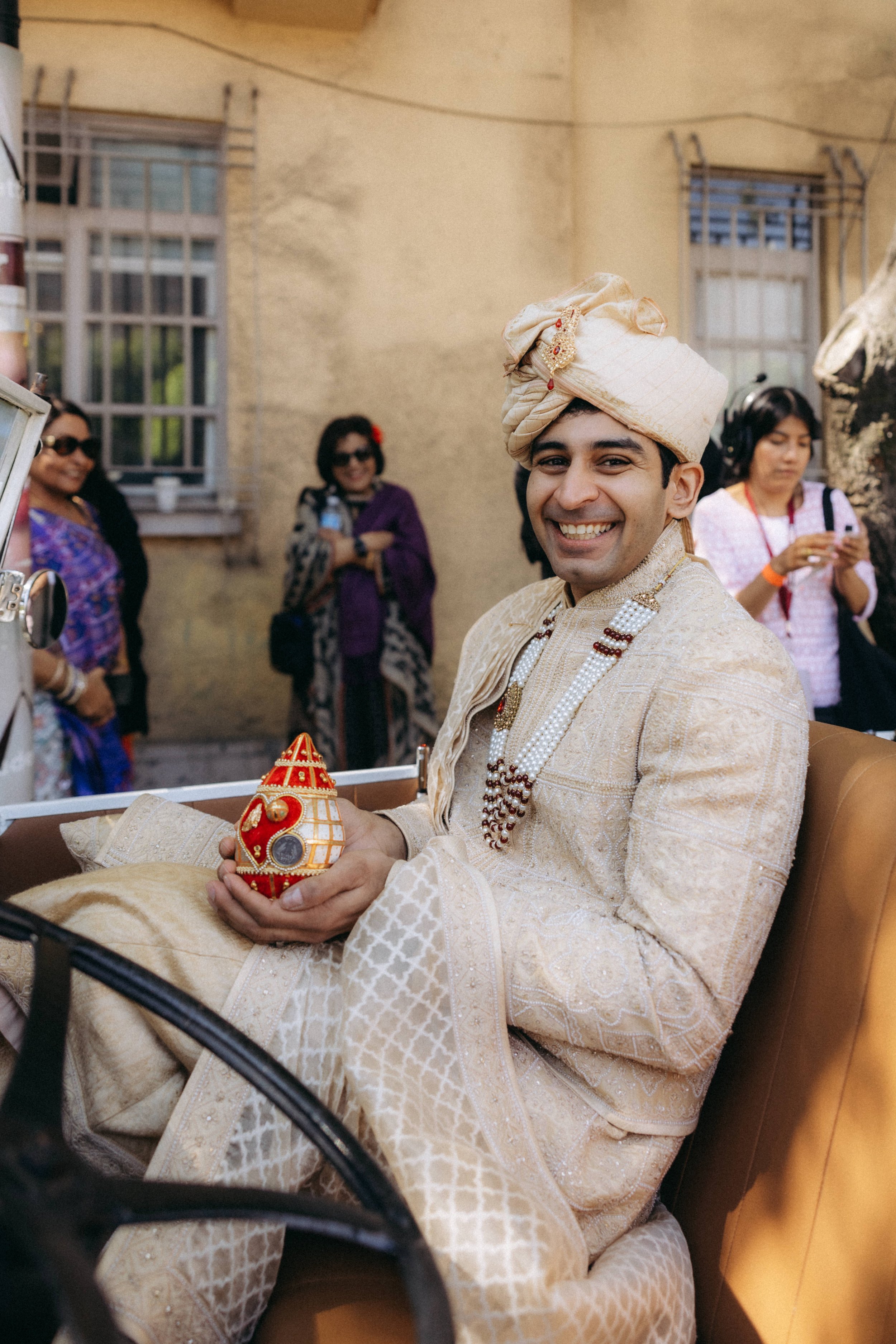 A smiling groom dressed in traditional Indian wedding attire, including an ornate cream-colored sherwani and turban, sitting in a carriage holding a decorative ornament. Several women in colorful sarees and outfits are standing in the background outs