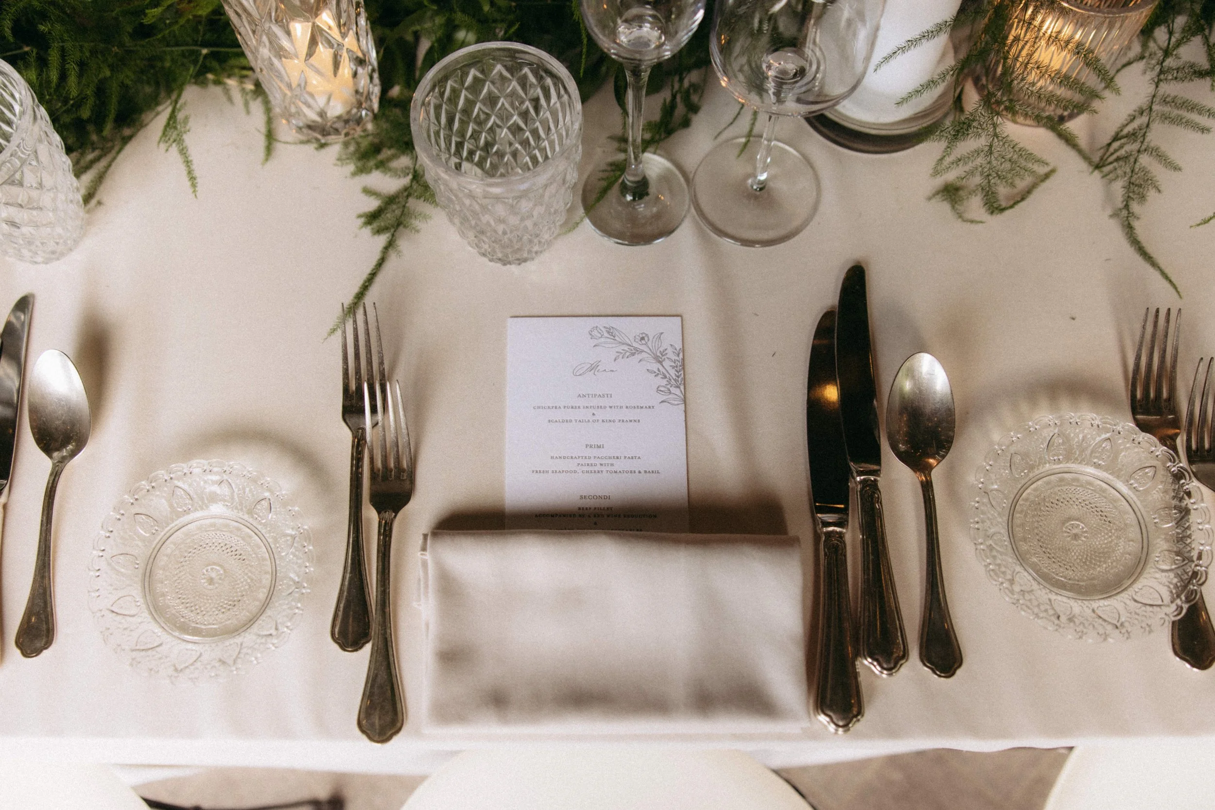 Table set for a meal, with clear glassware including wine glasses and water tumblers, elegant glass plates, silverware, and a menu card placed on a white tablecloth, with greenery and candles used as centerpieces.