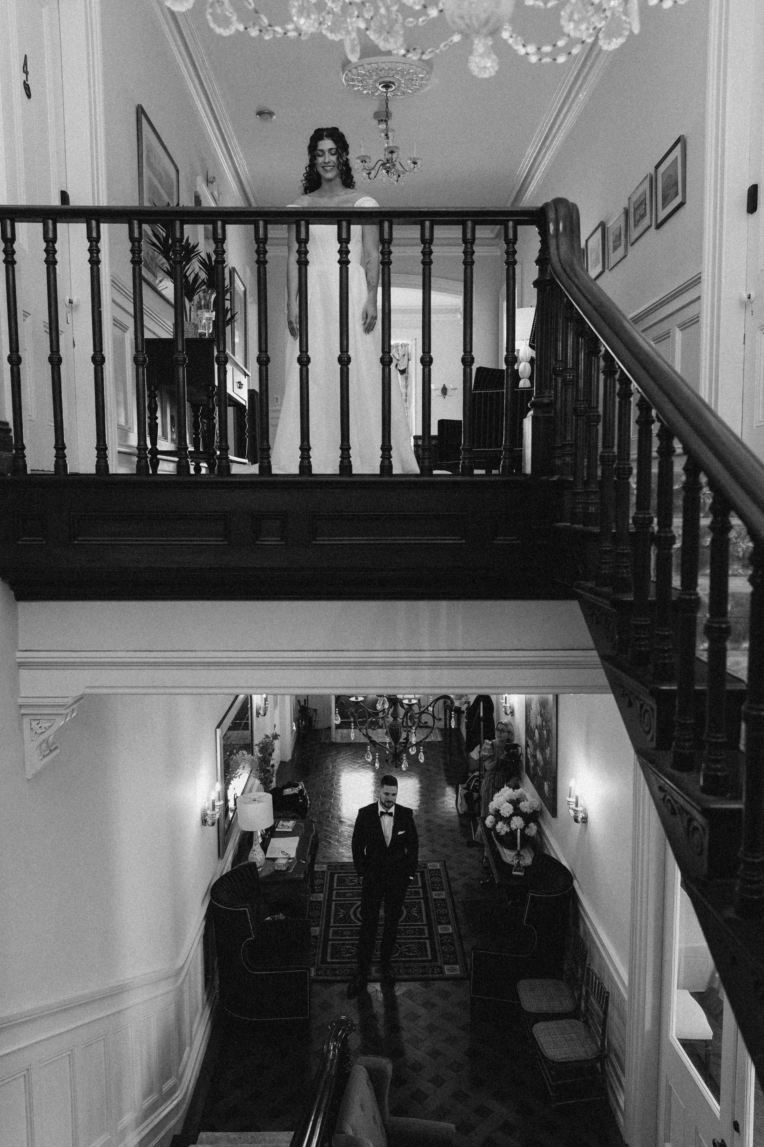 A woman in a wedding dress stands at the top of a staircase in a well-decorated home, smiling. Below, a man in a tuxedo stands in a hallway with elegant furniture, floral arrangements, and wall decor, smiling at the camera.