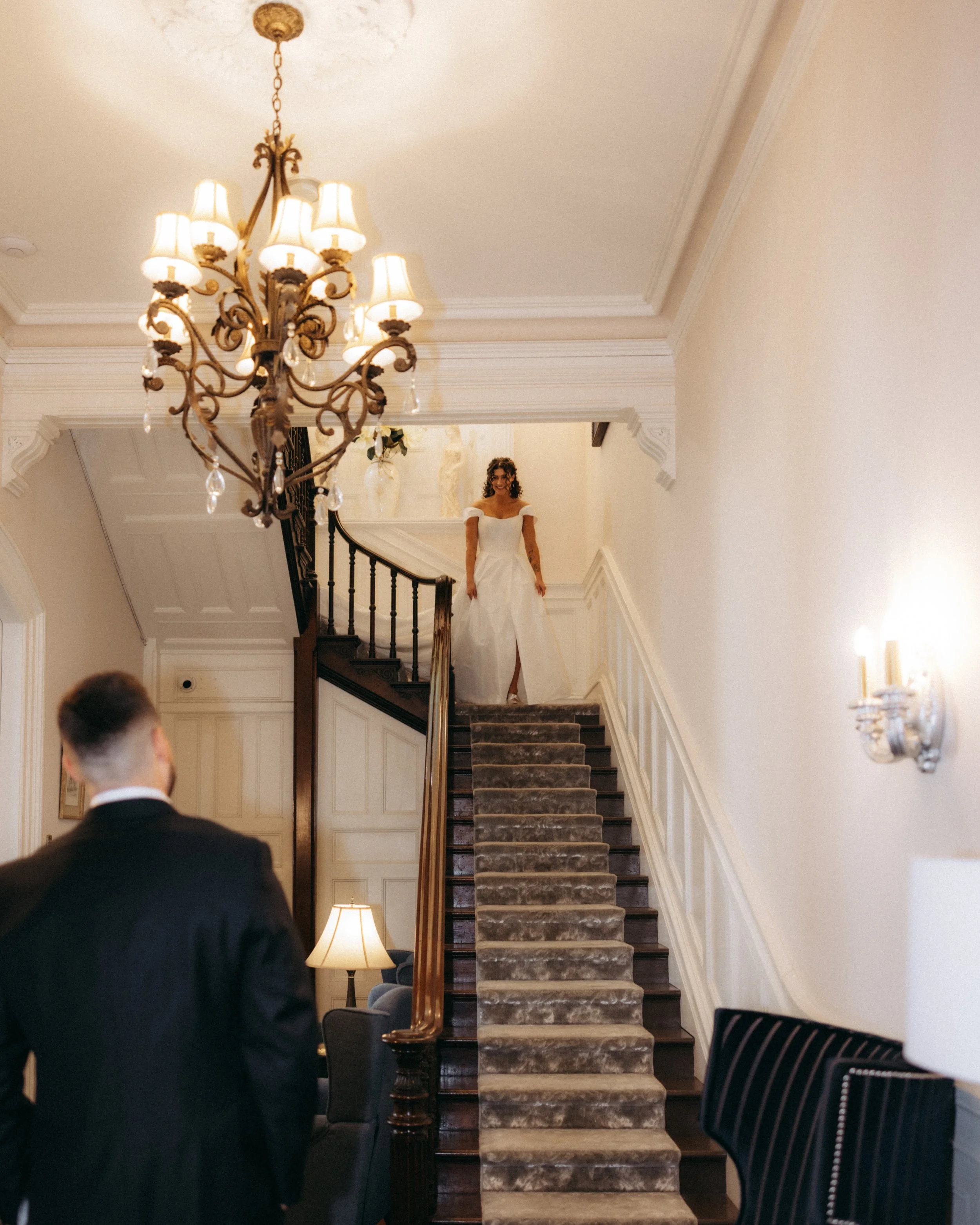 A woman in a wedding dress standing on a staircase in a decorated room, illuminated by a chandelier and lamps.