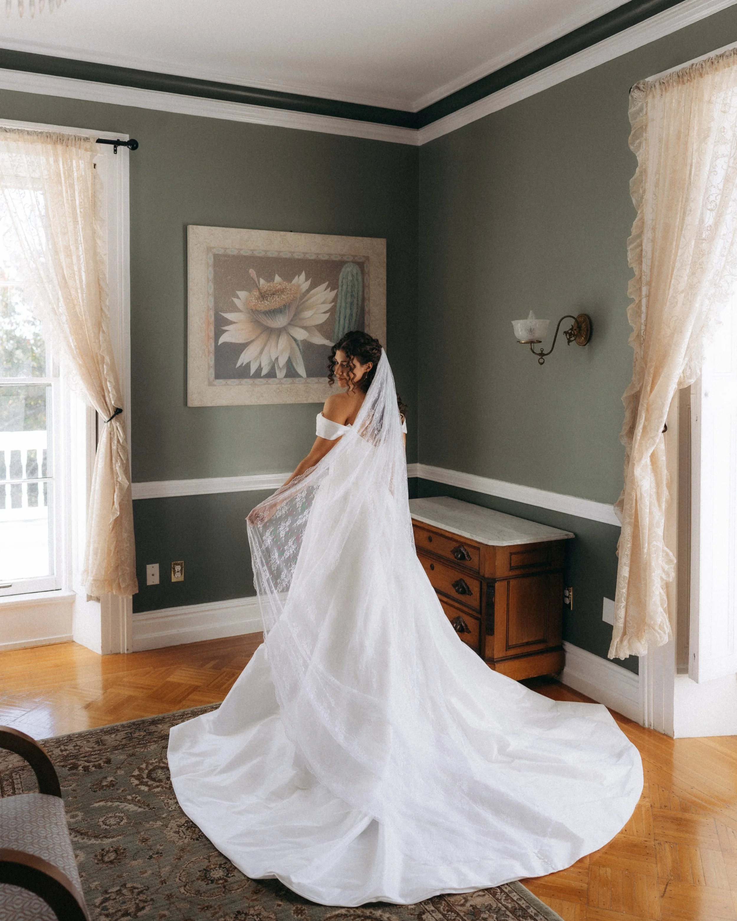 A woman in a white wedding dress with a veil standing in a room with dark green walls, cream-colored curtains, and art depicting a lotus flower and cactus on the wall.