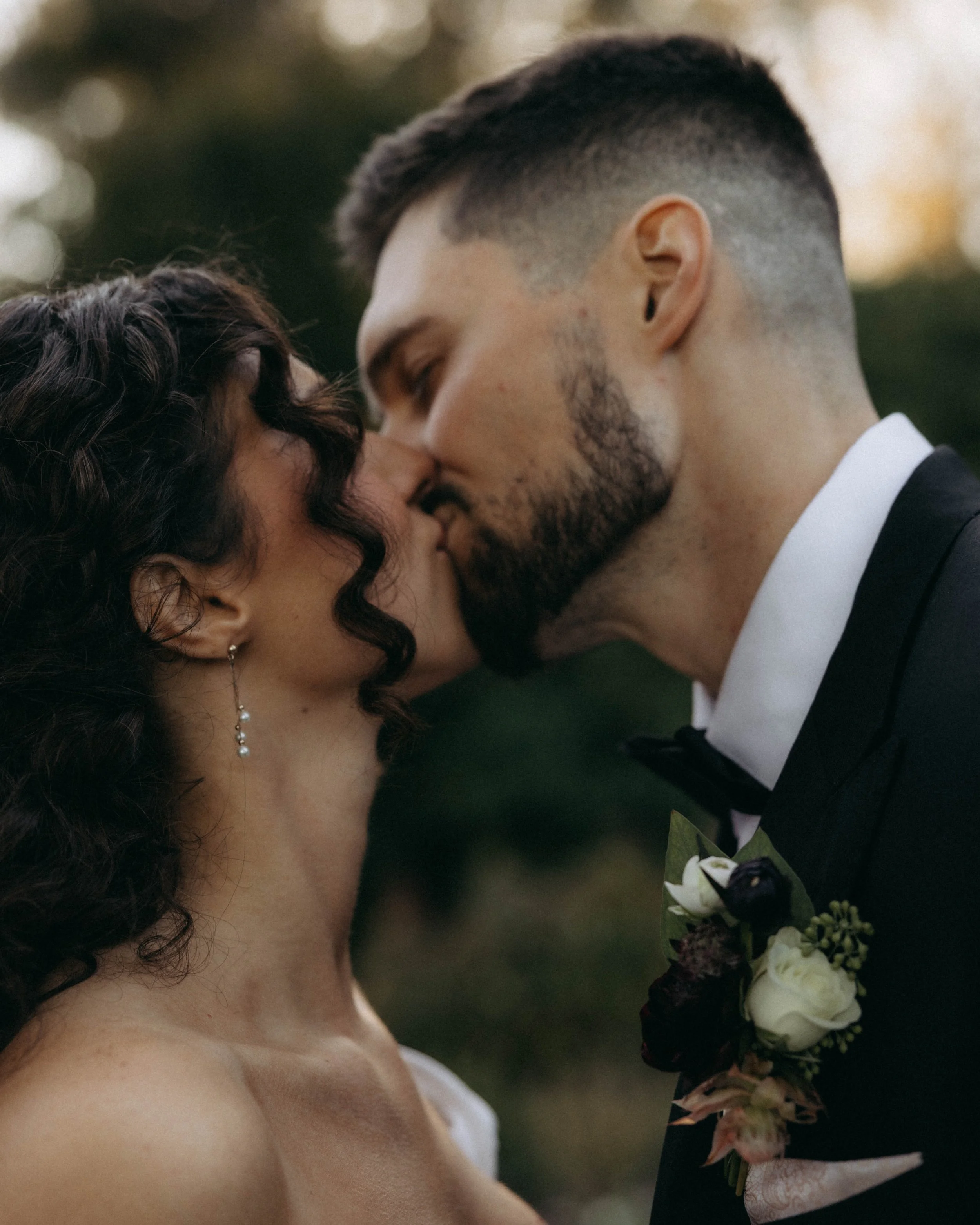 Close-up of a bride and groom sharing a gentle kiss outdoors, with the groom holding a bouquet of white and dark purple flowers.