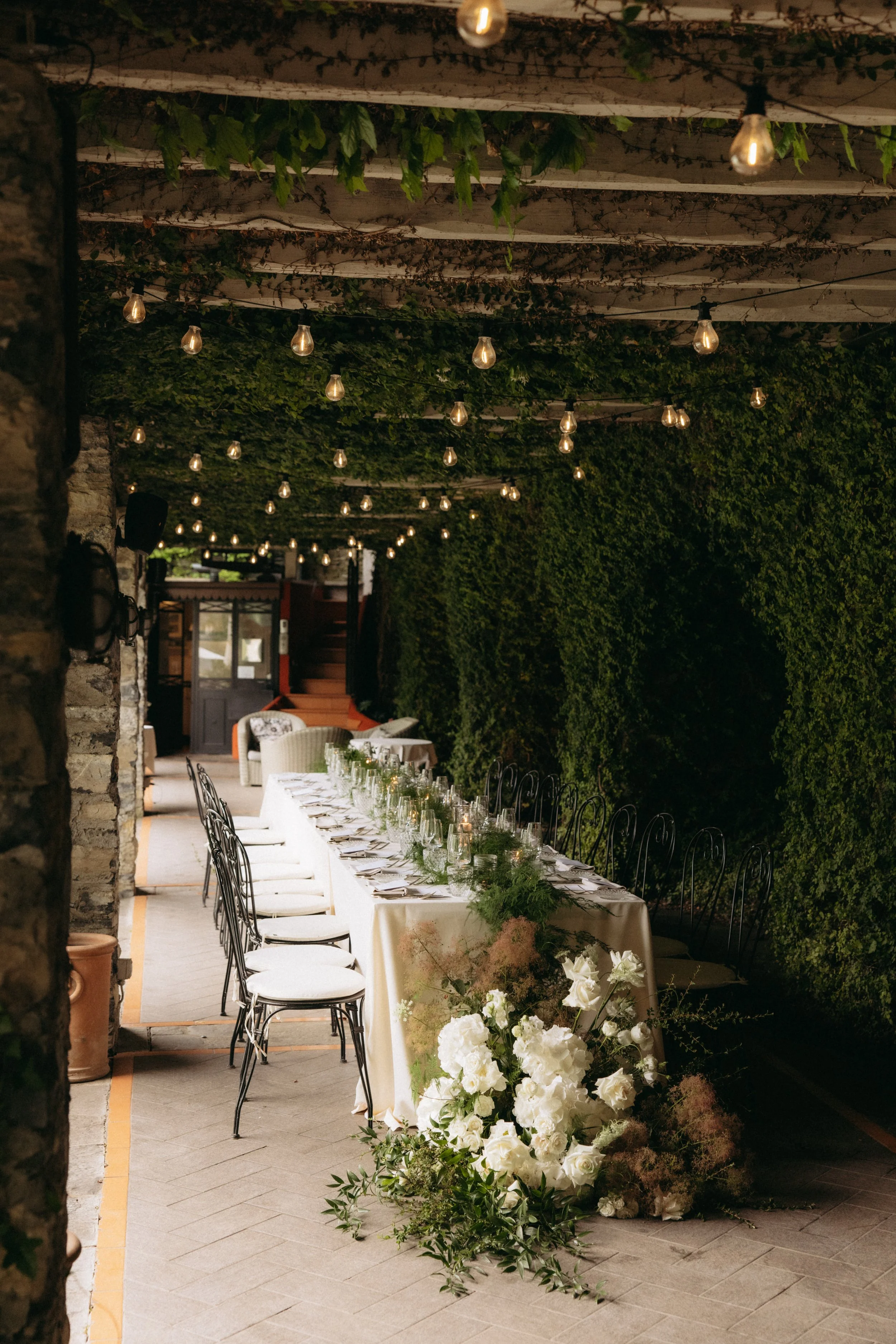 A decorated outdoor dining area with a long table covered with a white tablecloth, set with glassware and plates, surrounded by black chairs, and adorned with a floral centerpiece of white flowers and greenery. The area is under a pergola covered wit