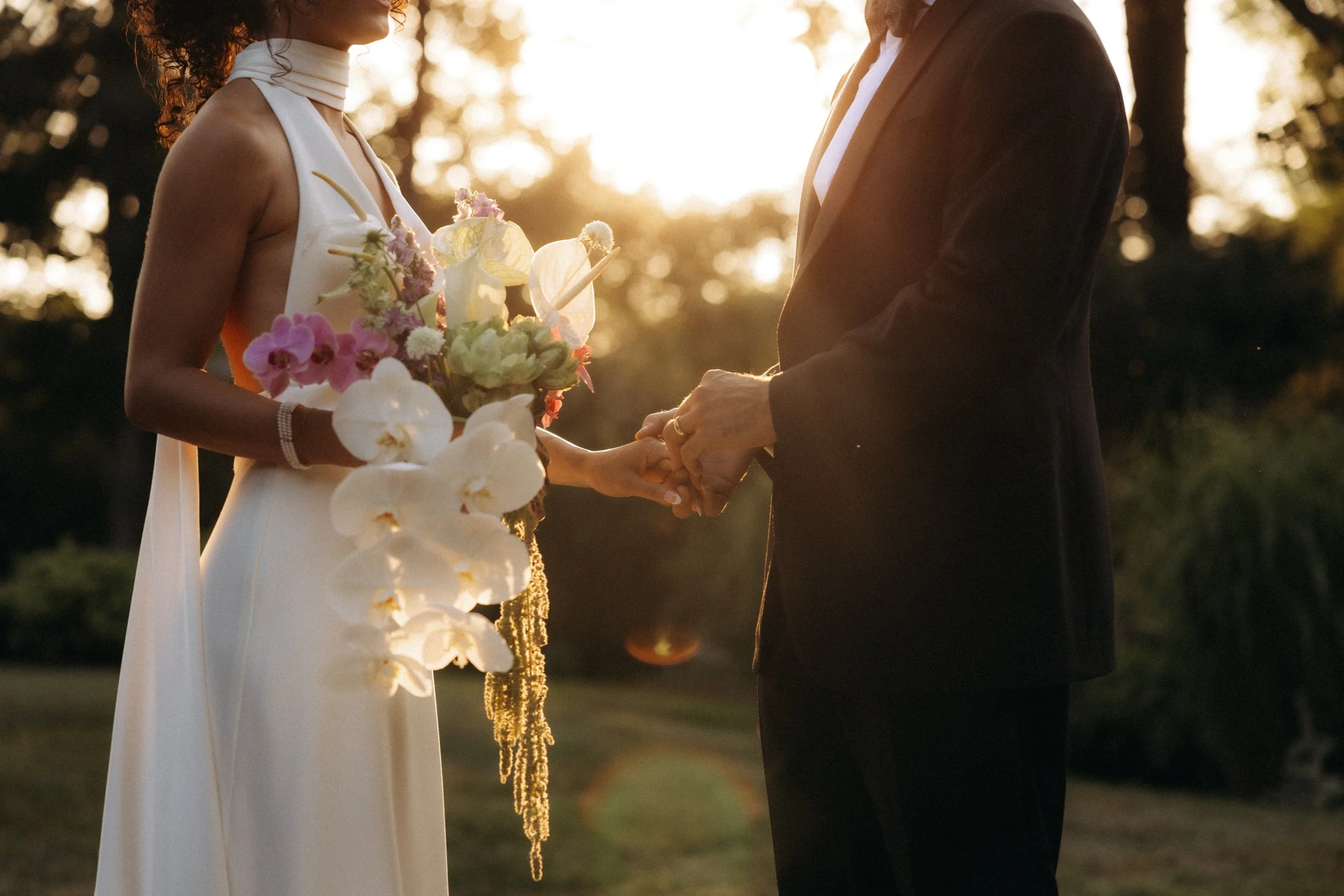 A couple holding hands during their wedding ceremony outdoors at sunset, with the bride wearing a sleeveless white dress and holding a bouquet of pink and white flowers, and the groom in a dark suit.
