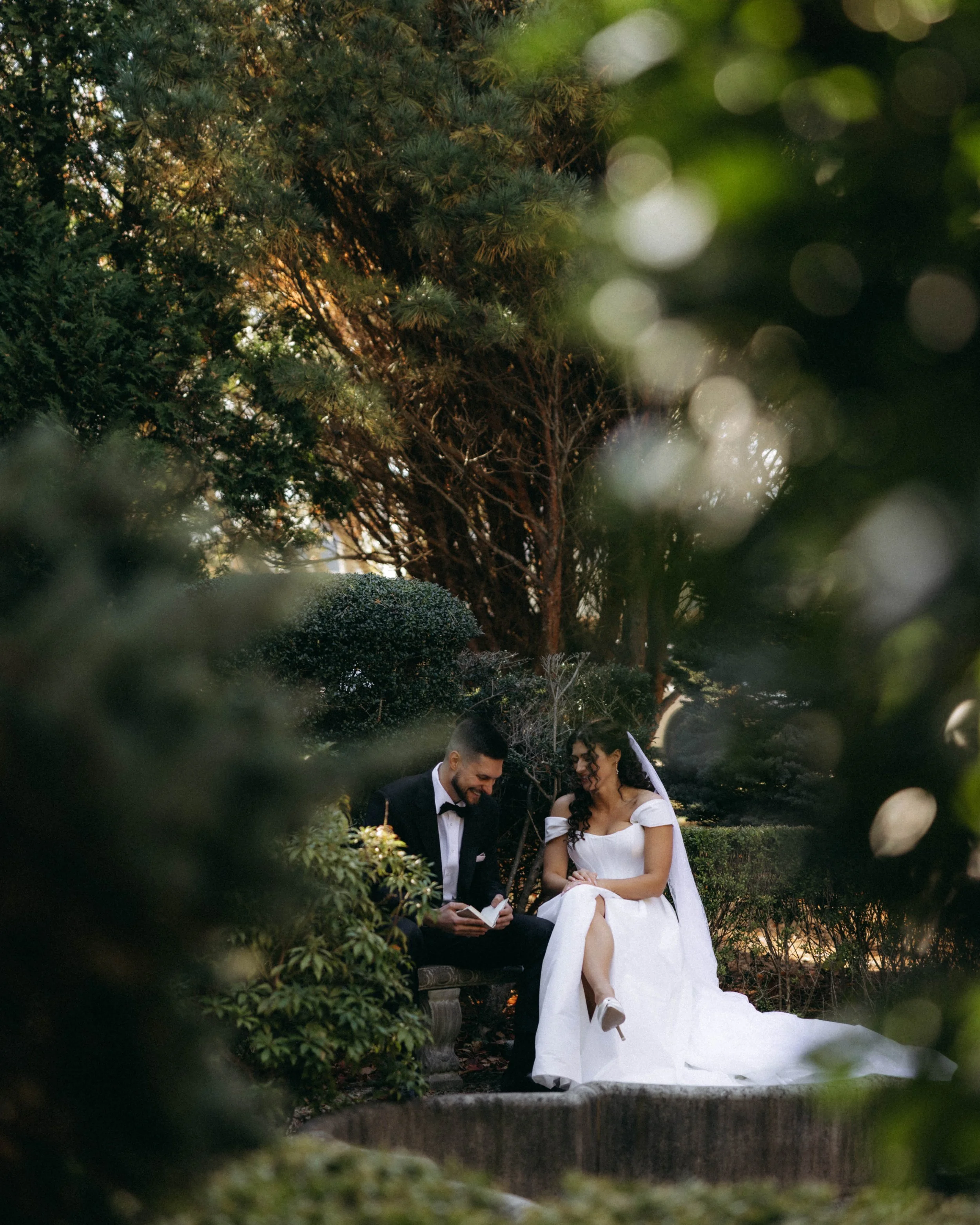 A bride and groom sitting on a park bench surrounded by greenery during a wedding ceremony, with the bride in a white dress and veil, and the groom in a black tuxedo, both smiling.