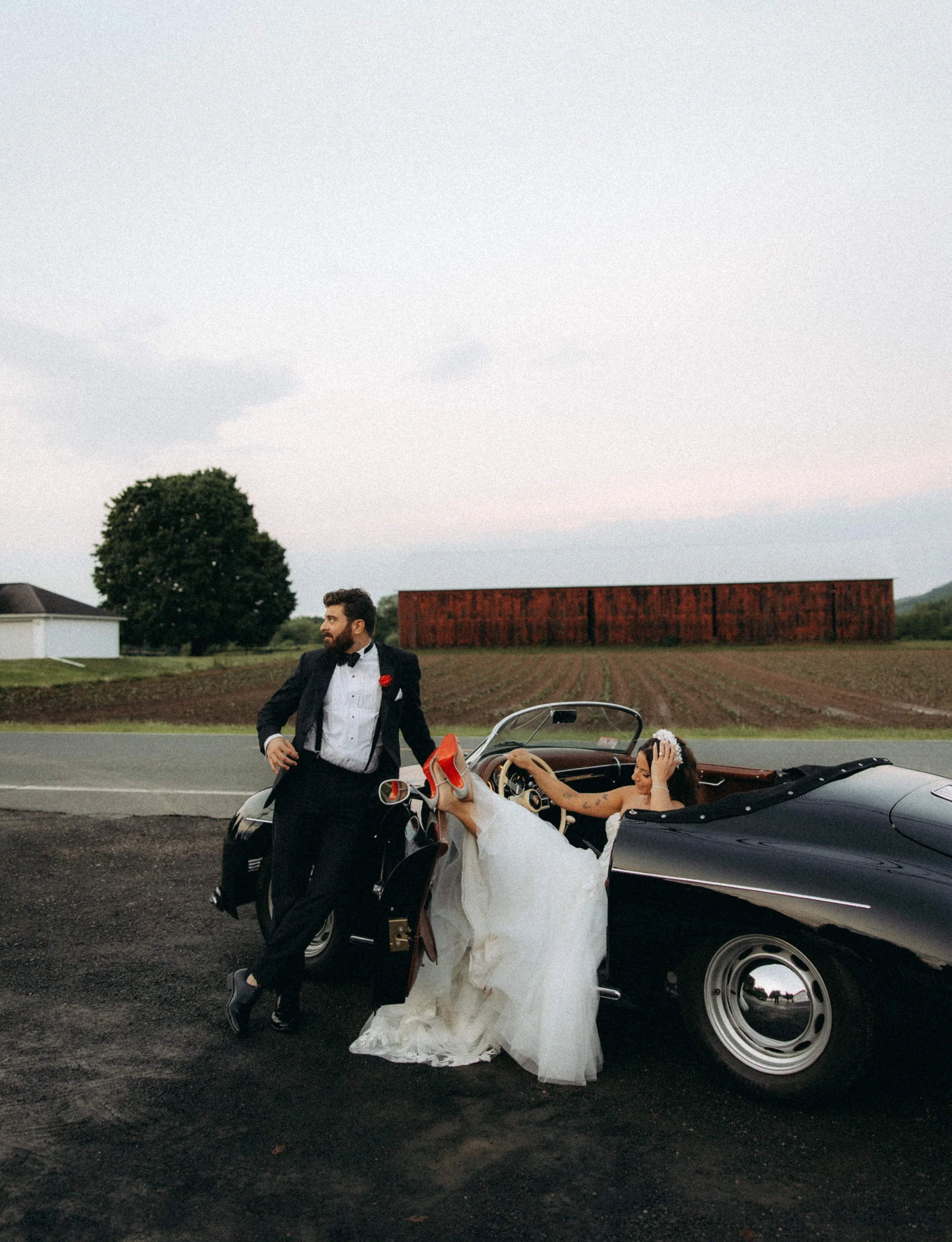 A man in a tuxedo and a woman in a wedding dress are next to a black convertible classic car on a rural road during the evening. The woman is sitting inside the car with her legs up on the door, smiling and touching her hair, while the man stands out