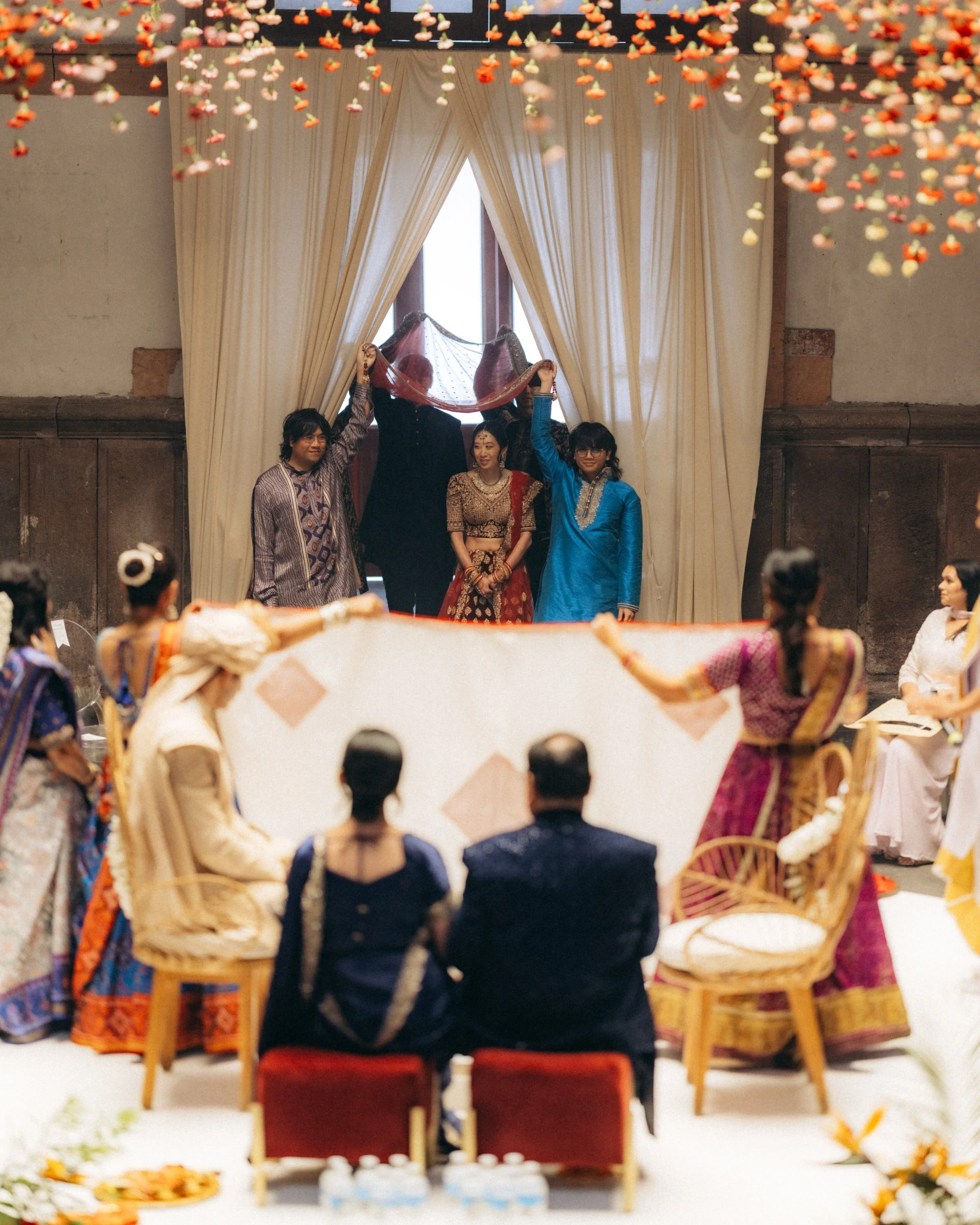 People in traditional Indian attire participating in a wedding ceremony in a decorated hall with cream curtains and hanging floral decorations.