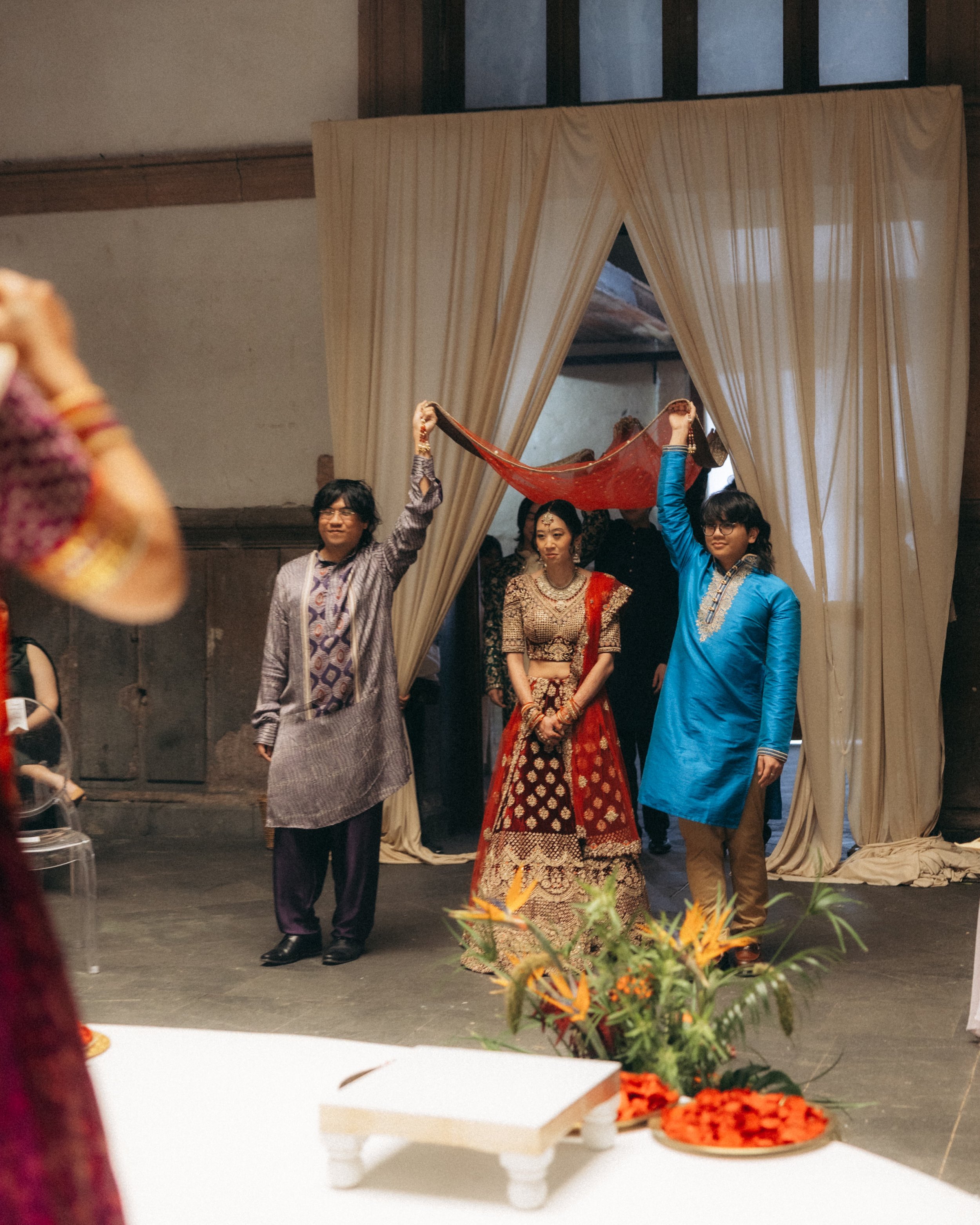 People participating in a traditional dance or ceremony at a wedding, with some holding a red cloth, standing in front of beige curtains, in a decorated indoor setting.