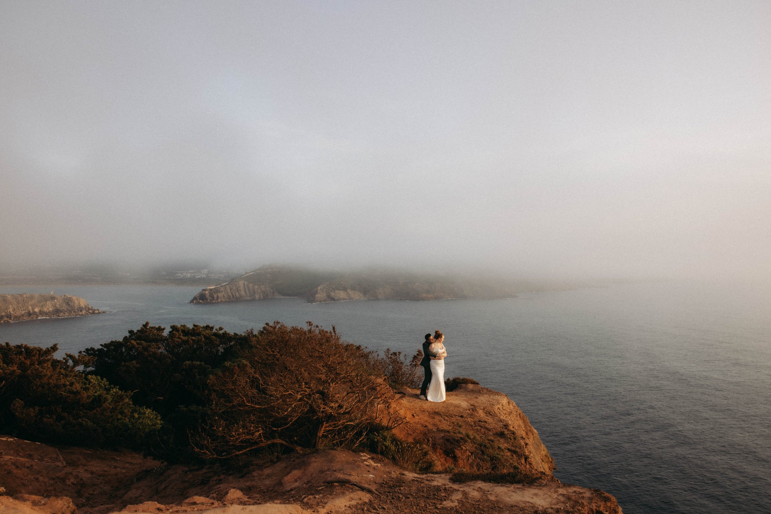 A bride and groom standing on a cliff overlooking the ocean with cliffs and hills in the background, during sunset or early evening.