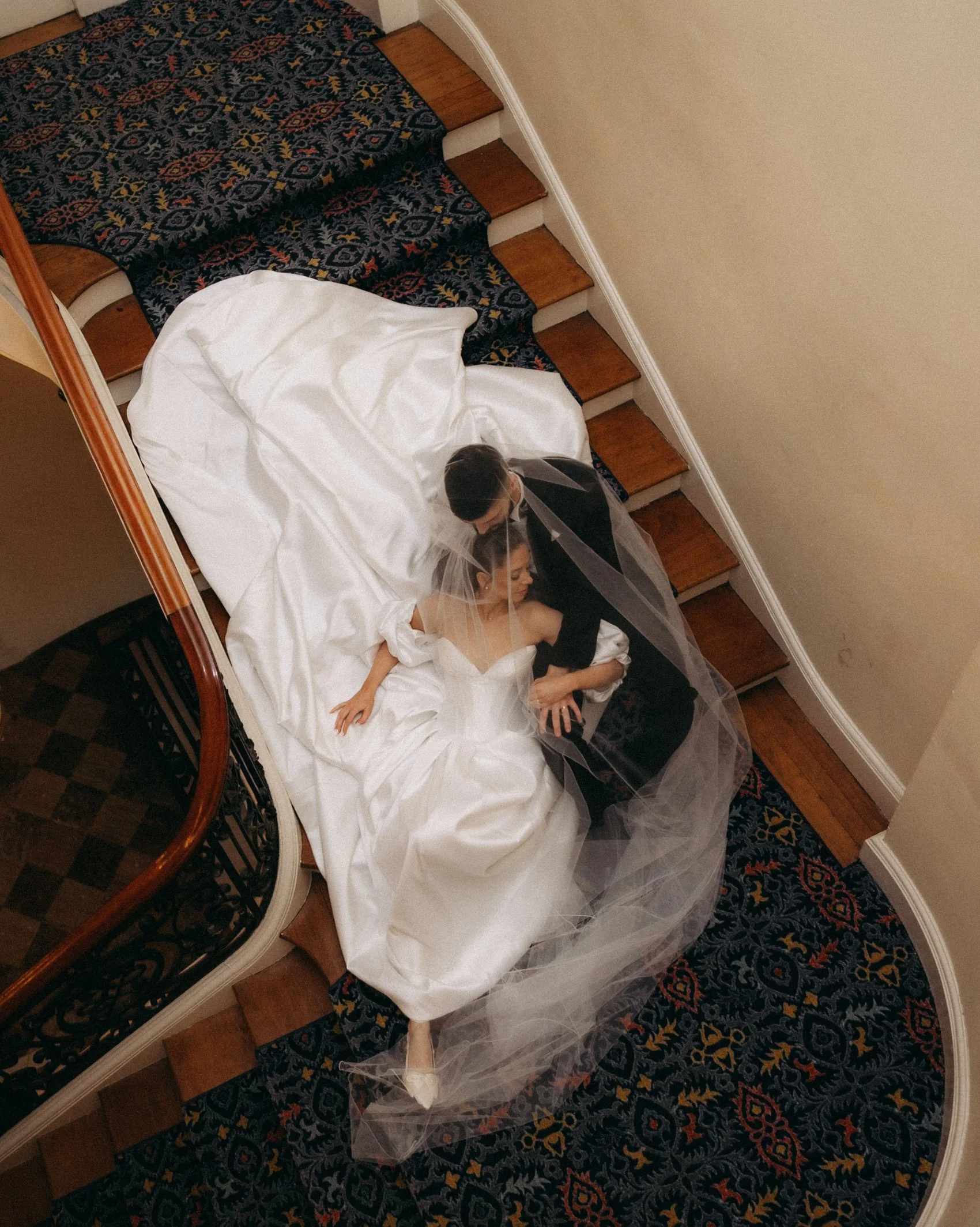 A newlywed couple on a wedding staircase, with the bride in a white wedding gown and veil, and the groom in a black tuxedo, standing close together in a hotel hallway with a patterned rug and wooden stairs.