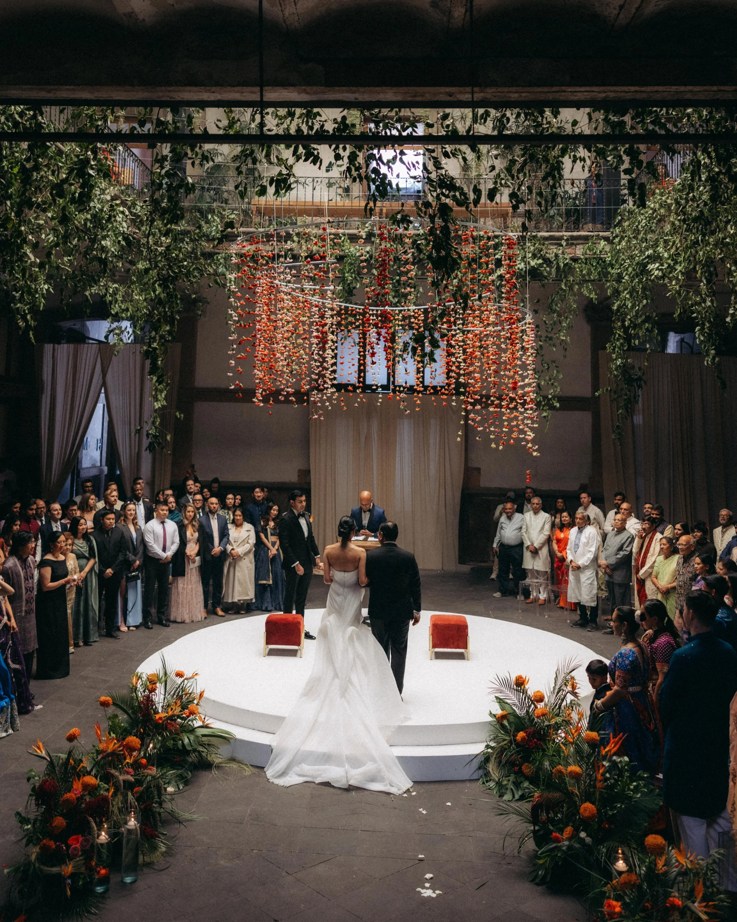 A wedding ceremony with a bride and groom standing on a circular platform, surrounded by guests. Floral arrangements and orange flowers decorate the area. A large chandelier made of hanging orange and white flowers hangs above. The venue has a high c