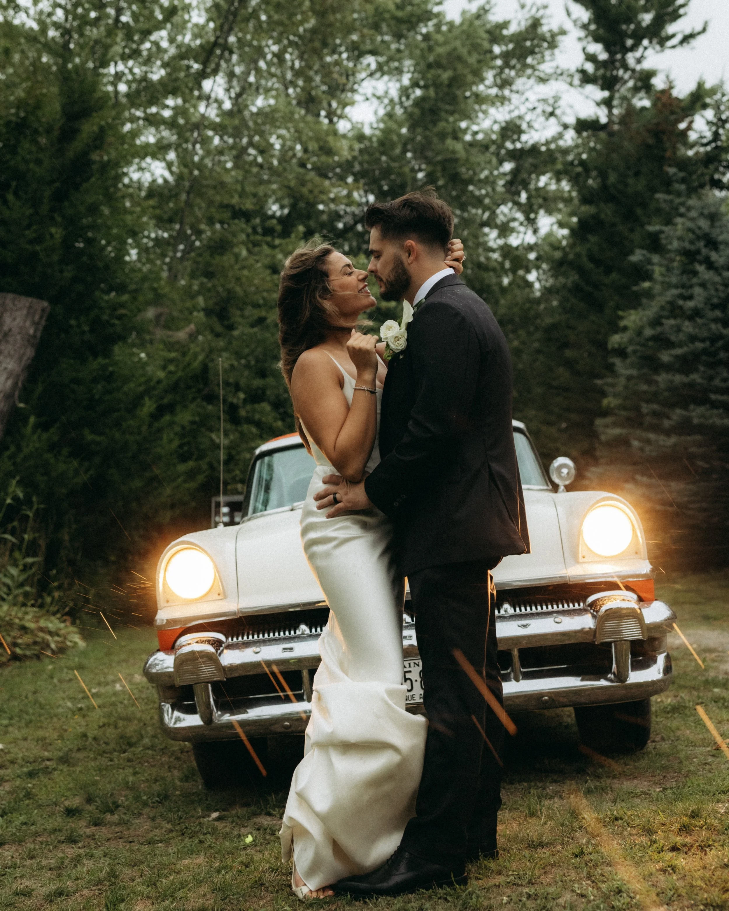 A couple dressed in wedding attire standing close together in front of a vintage white car. The woman is wearing a white wedding dress and the man is wearing a black tuxedo. They are outdoors amidst greenery, sharing an intimate moment with their fac