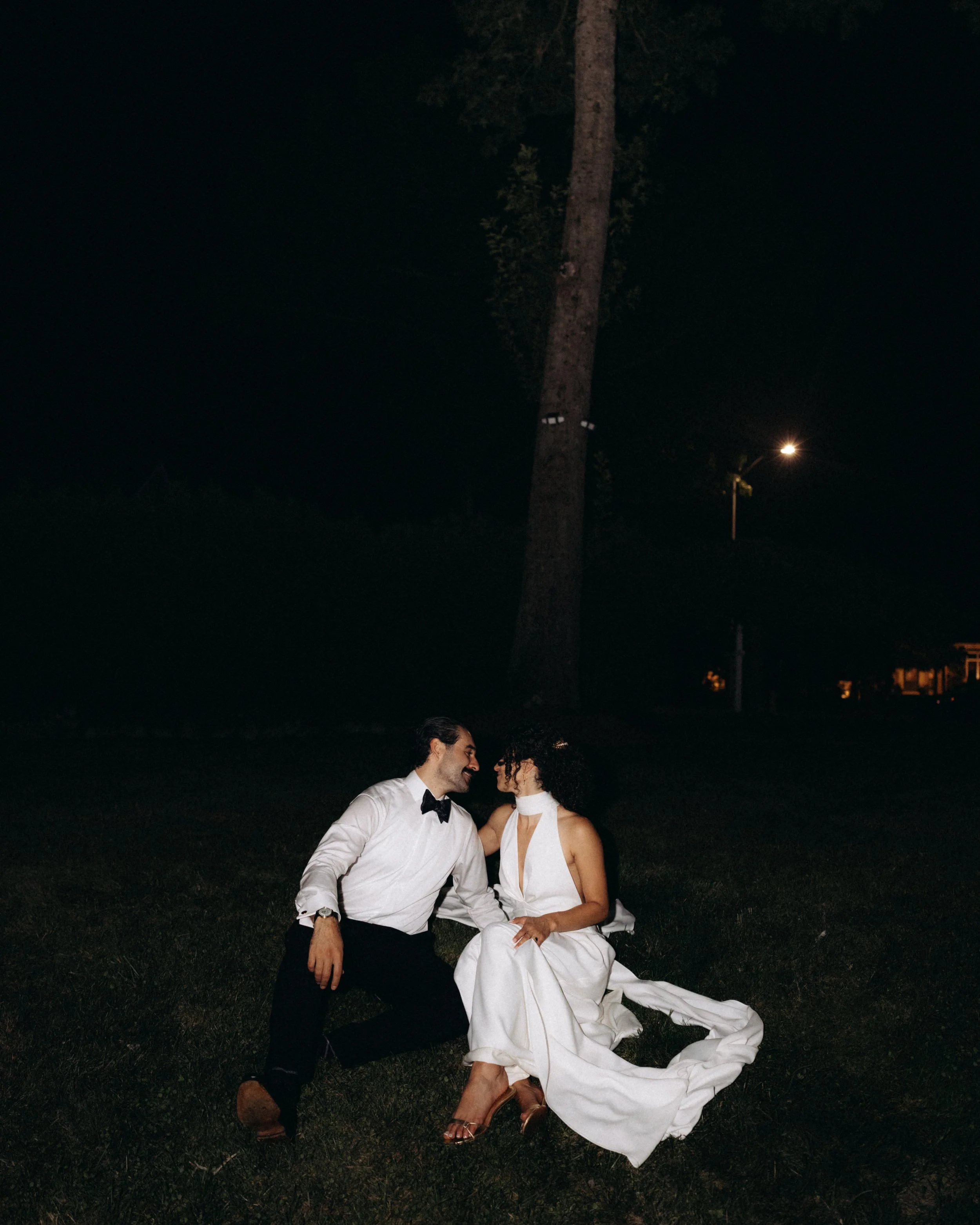 A couple dressed in formal attire, sitting outdoors at night, smiling and facing each other affectionately.