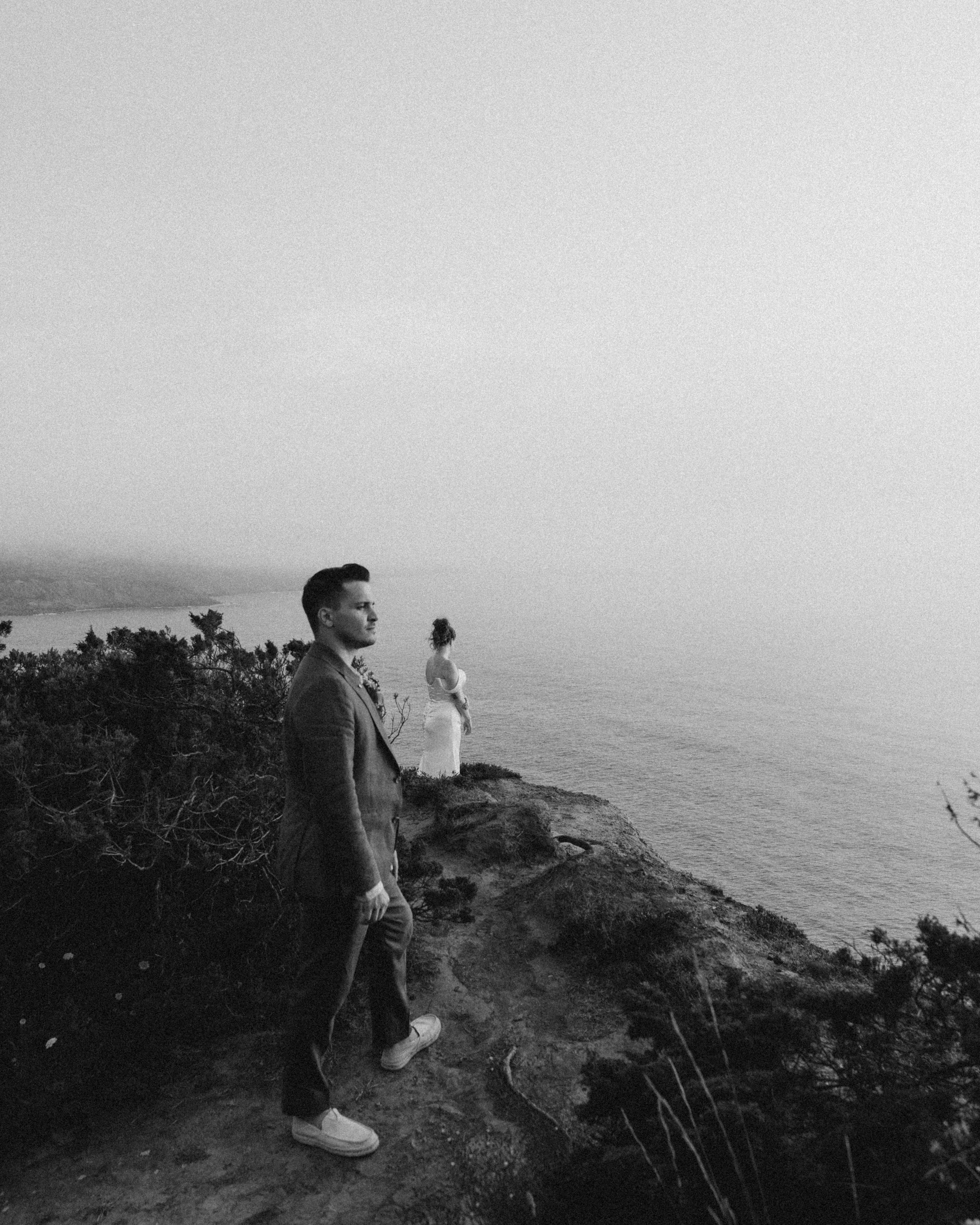 A black and white photo of three people standing on a rocky cliff overlooking the ocean. The person in the foreground is a young man wearing a jacket and sneakers, looking towards the left. Behind him, a woman in a dress with her hair tied up is look