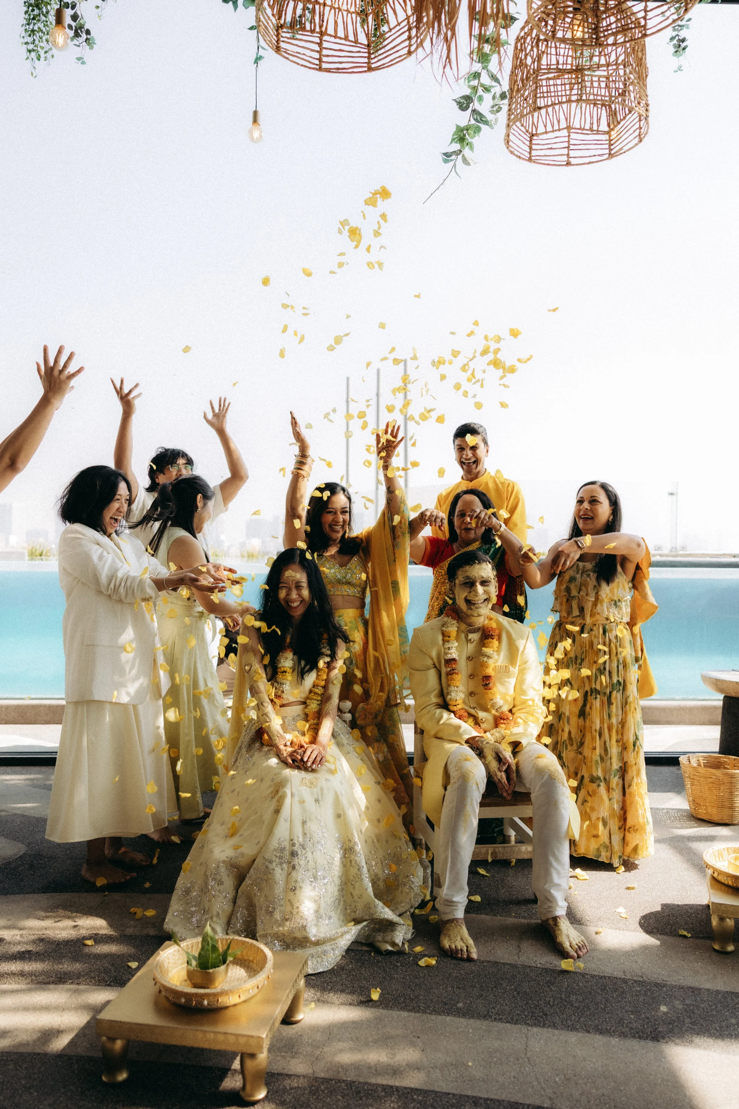 A group of people celebrating a traditional Indian wedding ceremony outdoors, with flower petals falling, some dressed in traditional Indian attire and others in formal wear, with a seated bride and groom in the center.