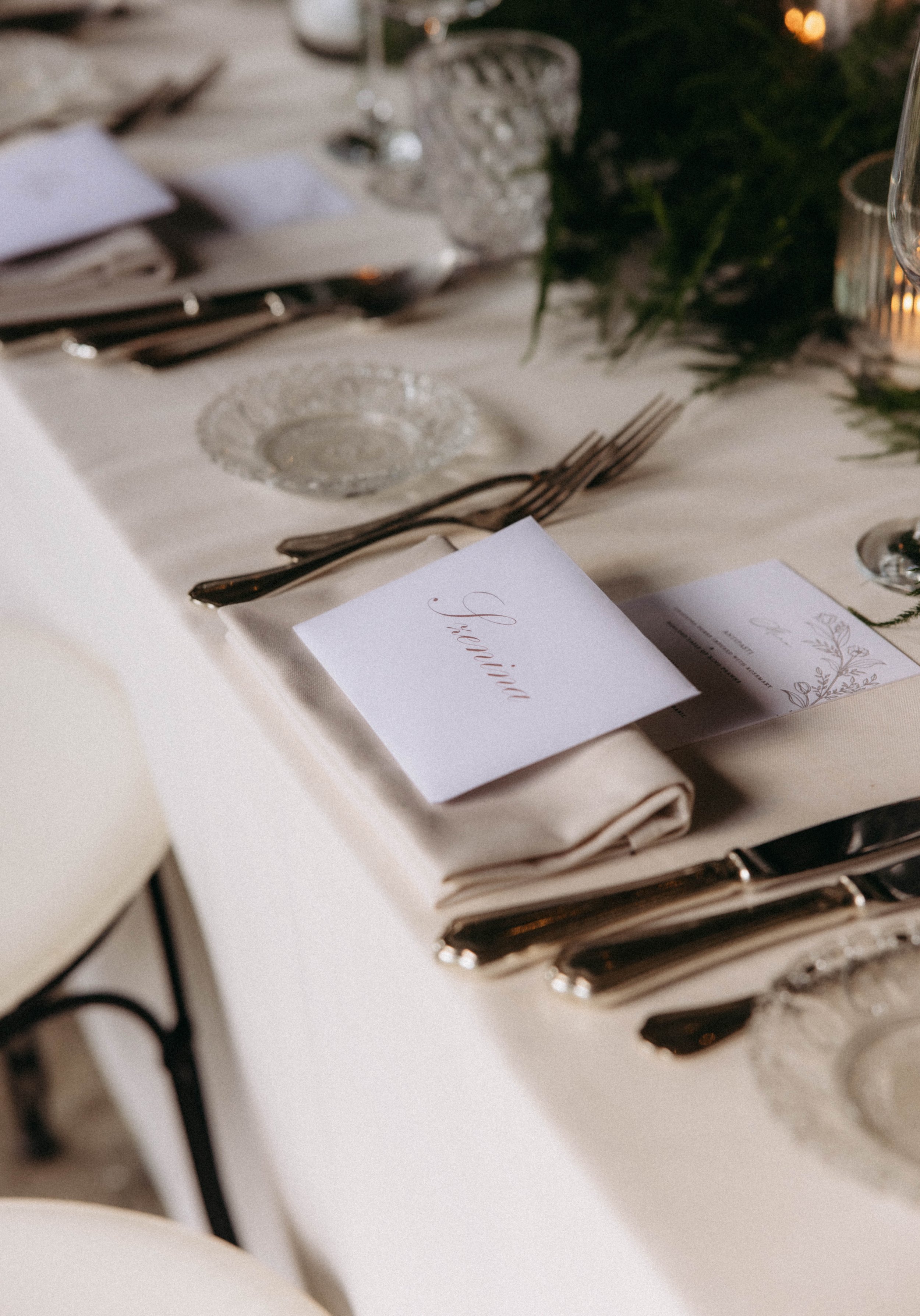 Elegant table setting with cutlery, glassware, a boxed MENU, and a garland centerpiece, suggesting a formal event or wedding reception.