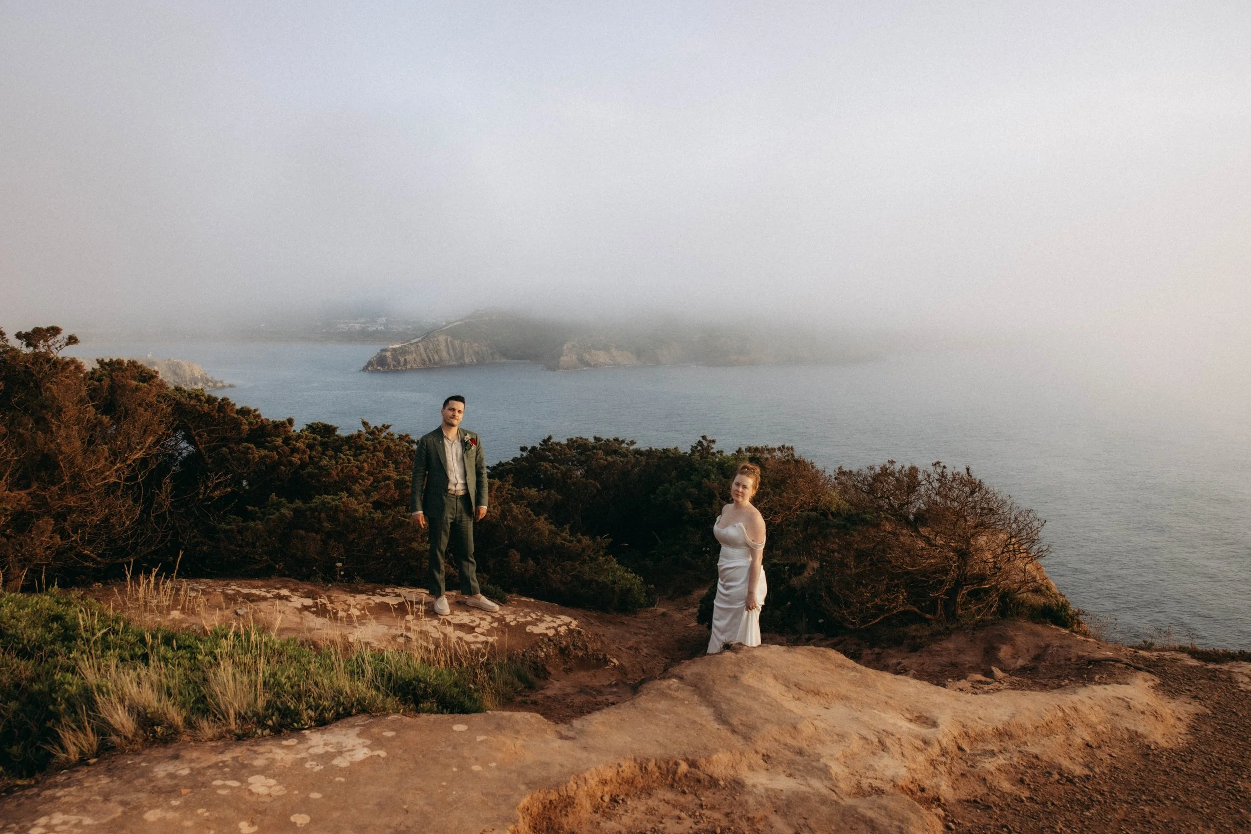 A couple dressed for a wedding standing on a rocky path surrounded by shrubs and trees, overlooking the ocean with a foggy hill in the background.