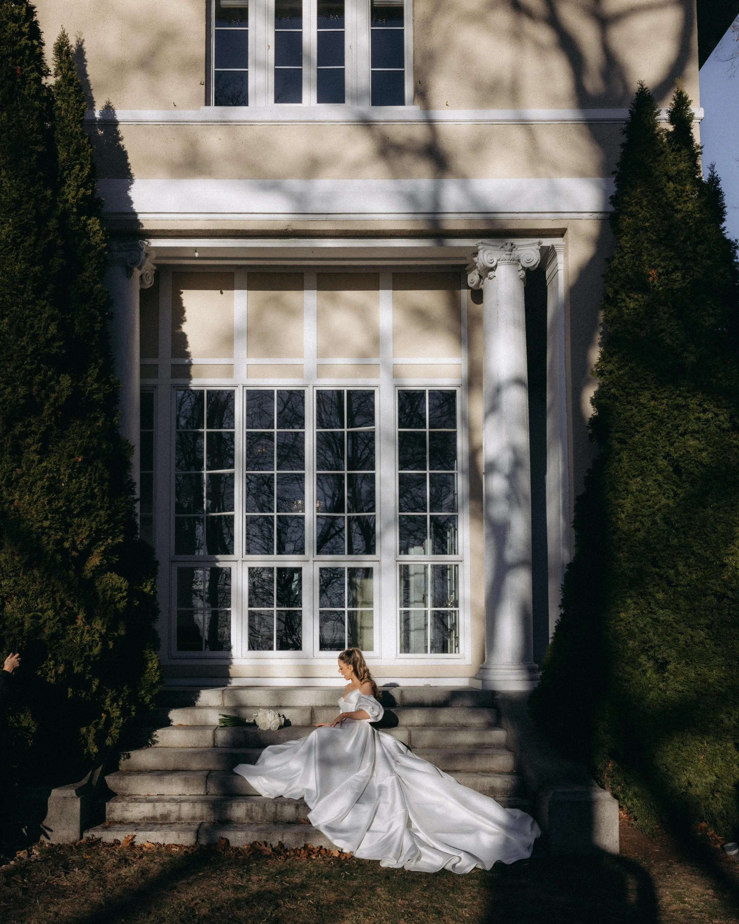A woman in a white wedding dress sits on stairs in front of a large building with classical columns and large windows, surrounded by tall trees casting shadows.