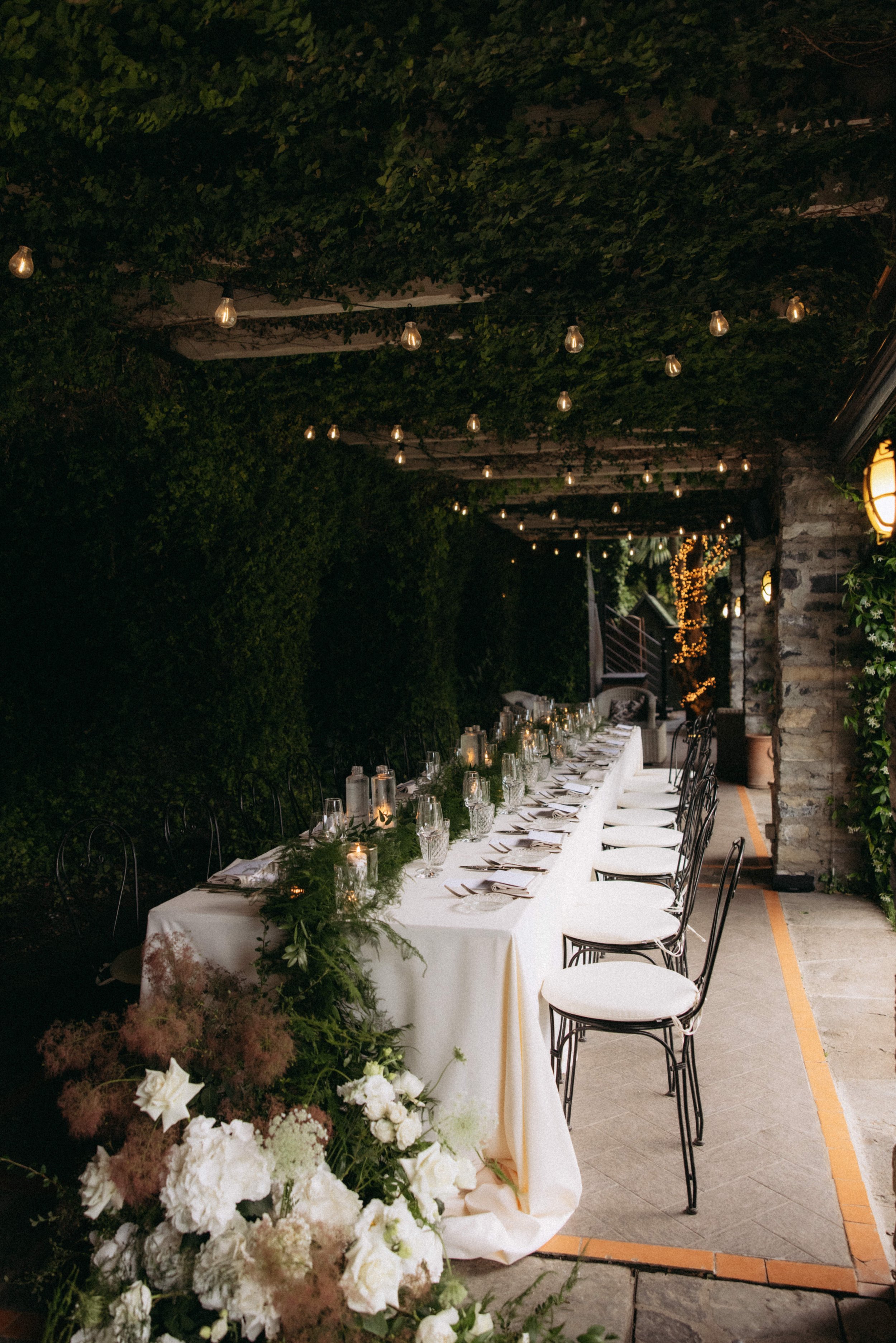 A long banquet table set for an outdoor evening event, decorated with white tablecloth, candles, and greenery, under a ceiling of string lights and surrounded by lush greenery and a stone wall.