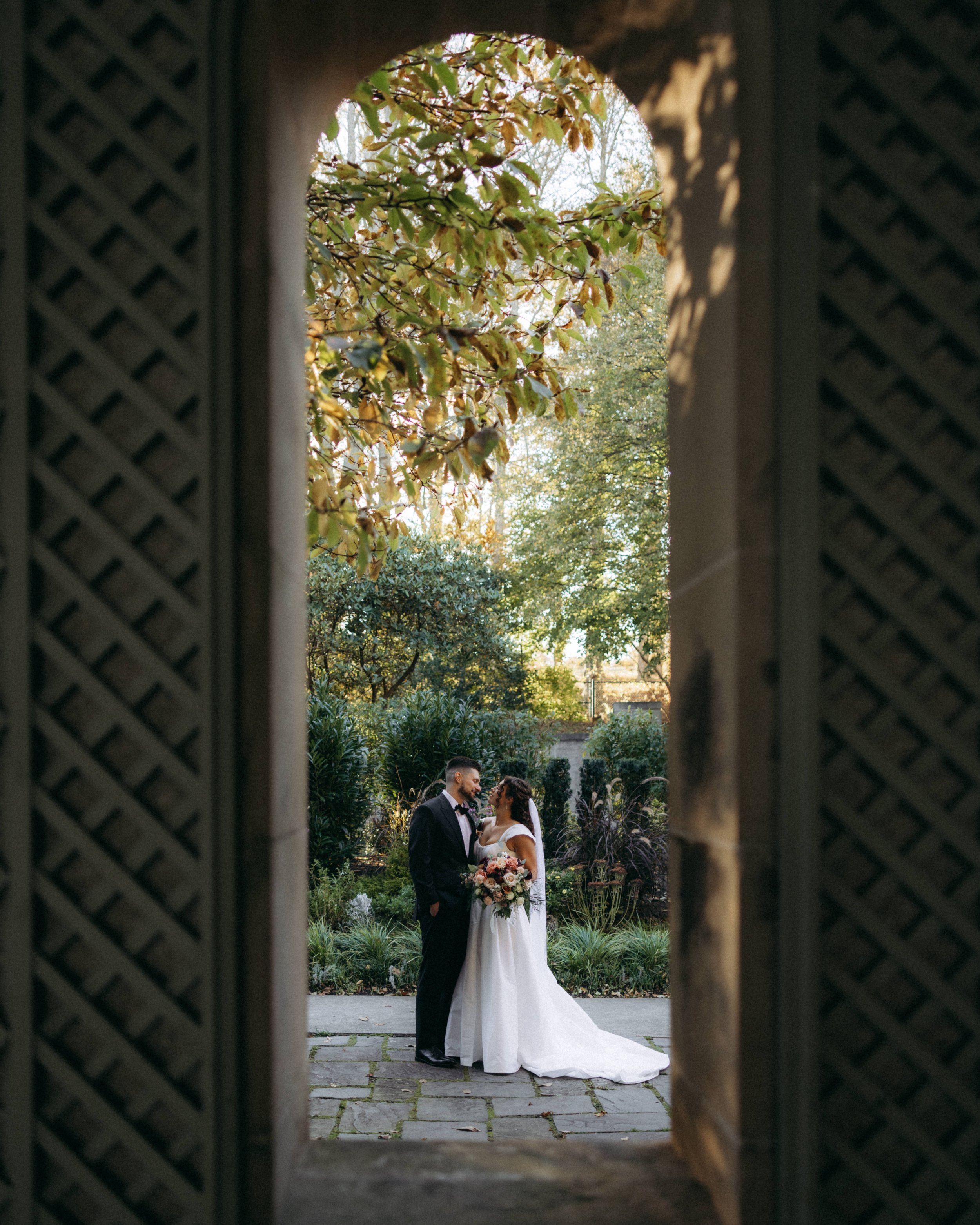 A bride and groom standing outdoors in a garden, seen through a decorative opening in a wooden fence or structure. The bride is holding a bouquet of flowers and is wearing a white wedding gown, while the groom is dressed in a black tuxedo. They are f