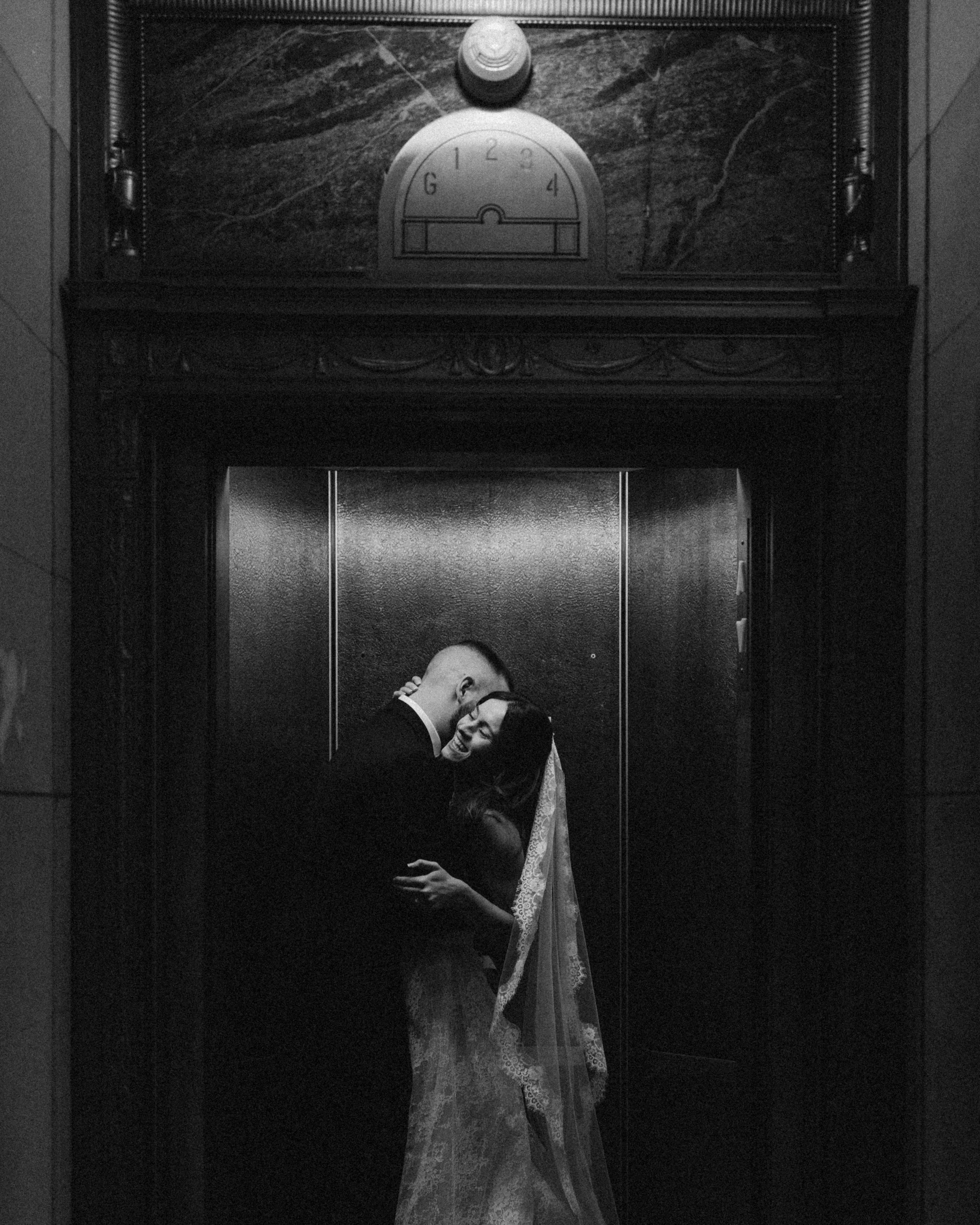 A couple sharing a kiss in an elevator, dressed in formal attire. The man is in a tuxedo and the woman in a lace wedding dress, holding each other close with their heads touching.