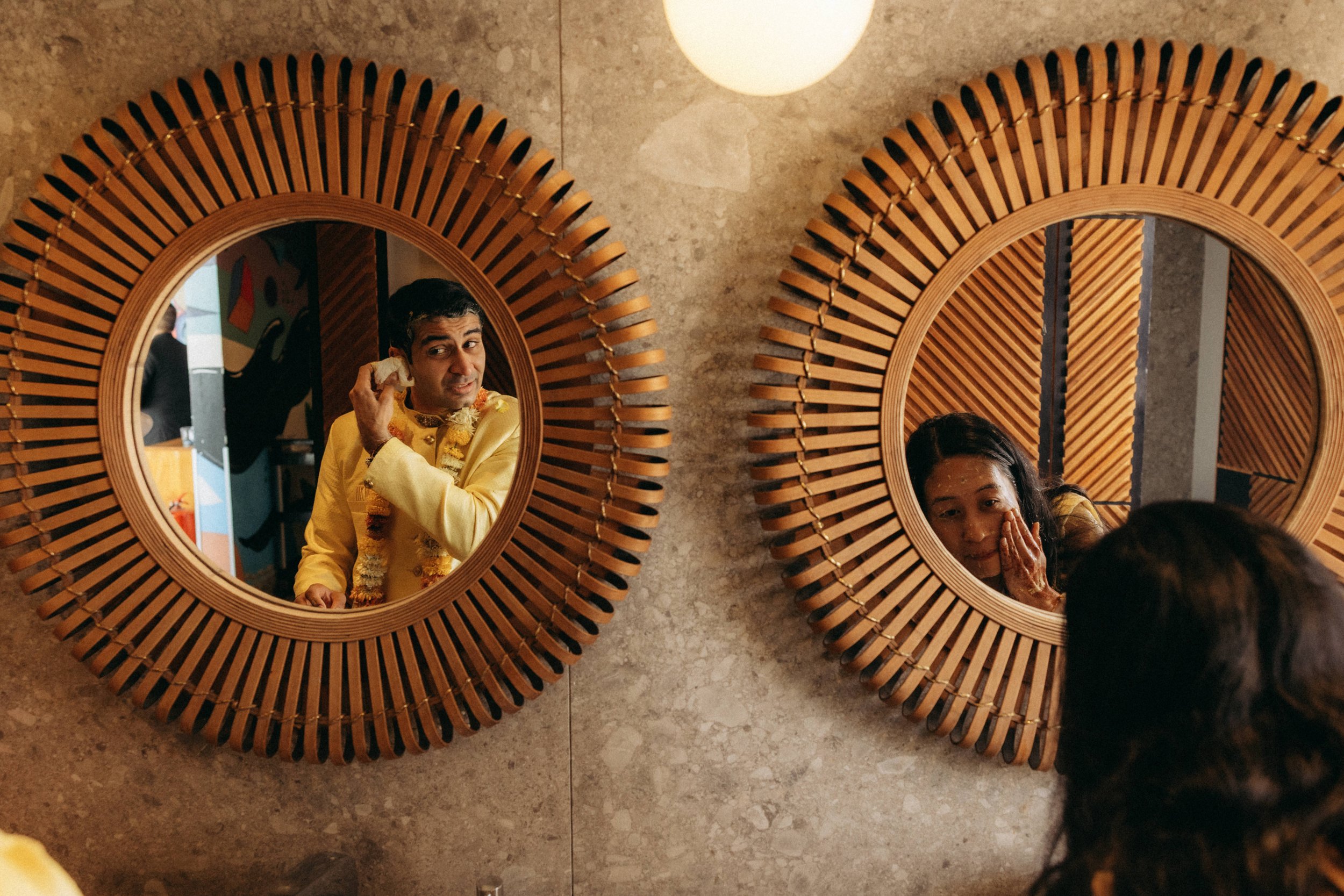 Two people looking into round mirrors with wooden frames mounted on a stone wall, one man in a yellow shirt holding an object near his ear and a woman resting her chin on her hand.