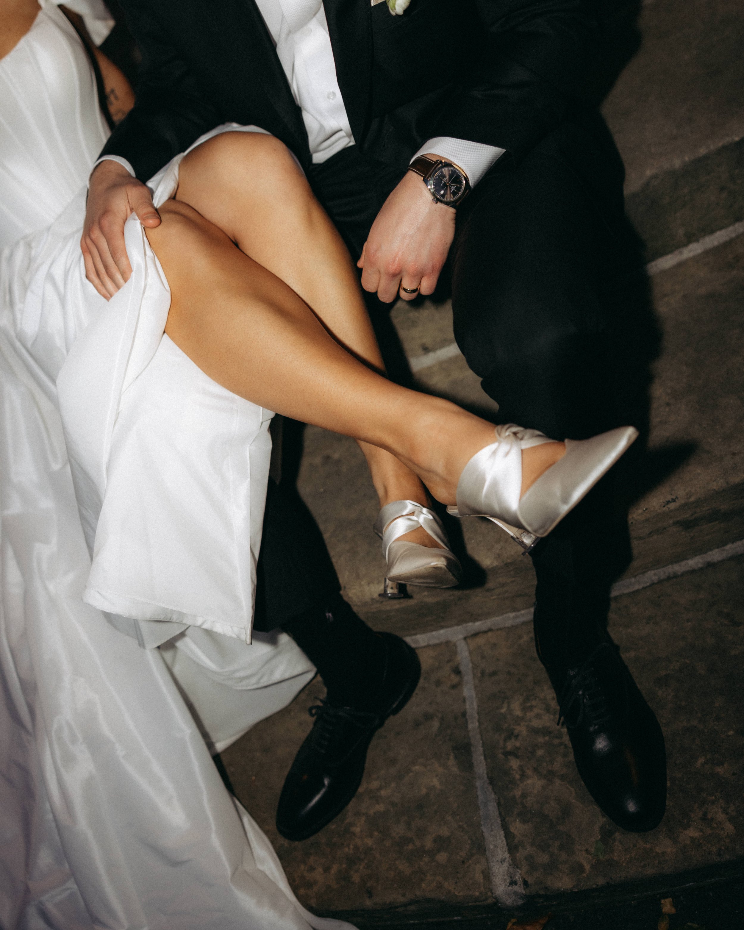 A man wearing a black suit and a white dress shirt sits with his legs crossed. His right hand rests on his knee, wearing a wristwatch with a dark face and leather strap. A woman in a white dress with satin shoes is sitting beside him, her right leg c