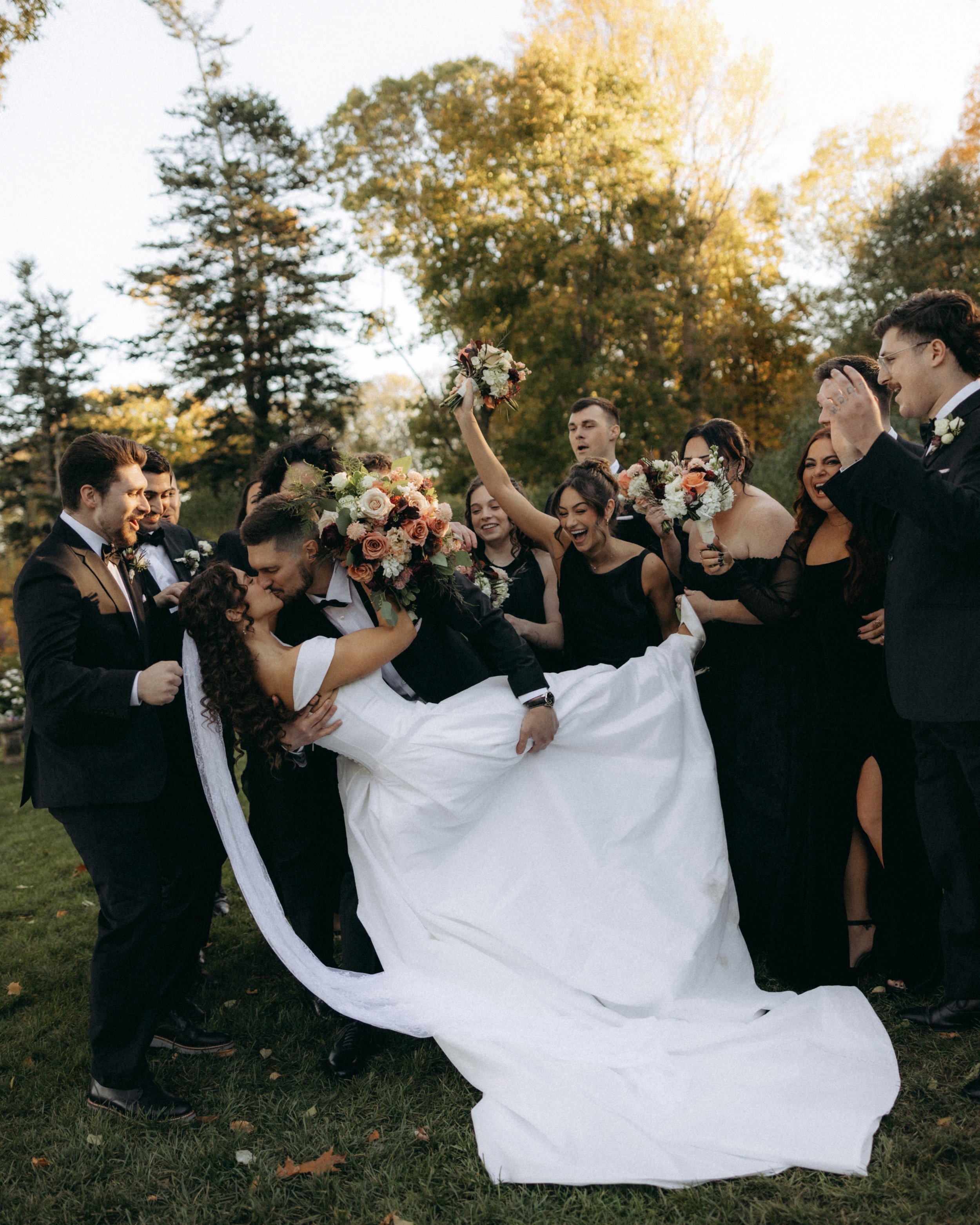 A group of people dressed in formal wedding attire celebrating outdoors, with an autumn backdrop of trees with yellow and orange leaves. The bride is being lifted, holding a bouquet, and others around her cheer and smile.