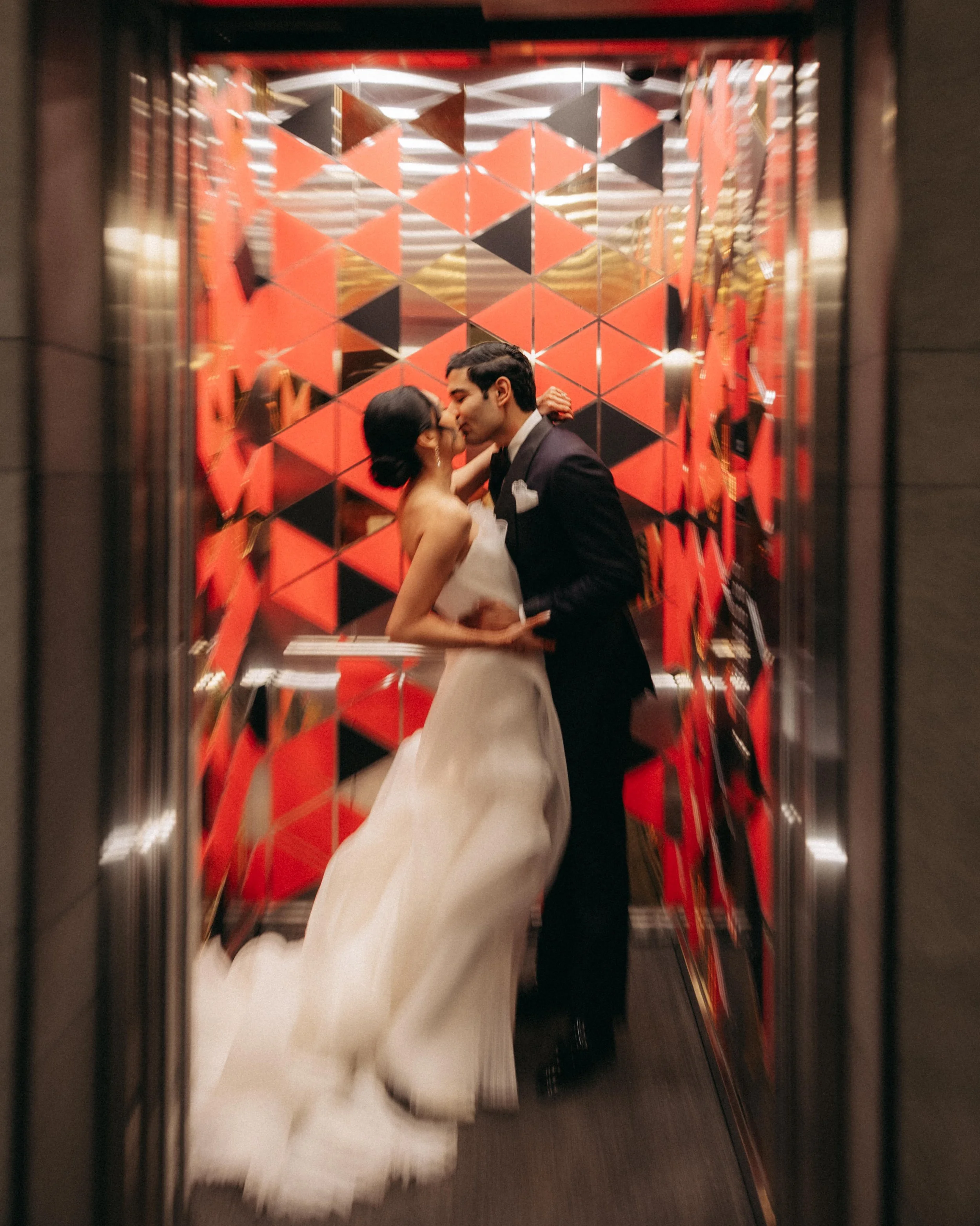 Couple in wedding attire sharing a kiss inside an elevator with gold and red mirrored panels.