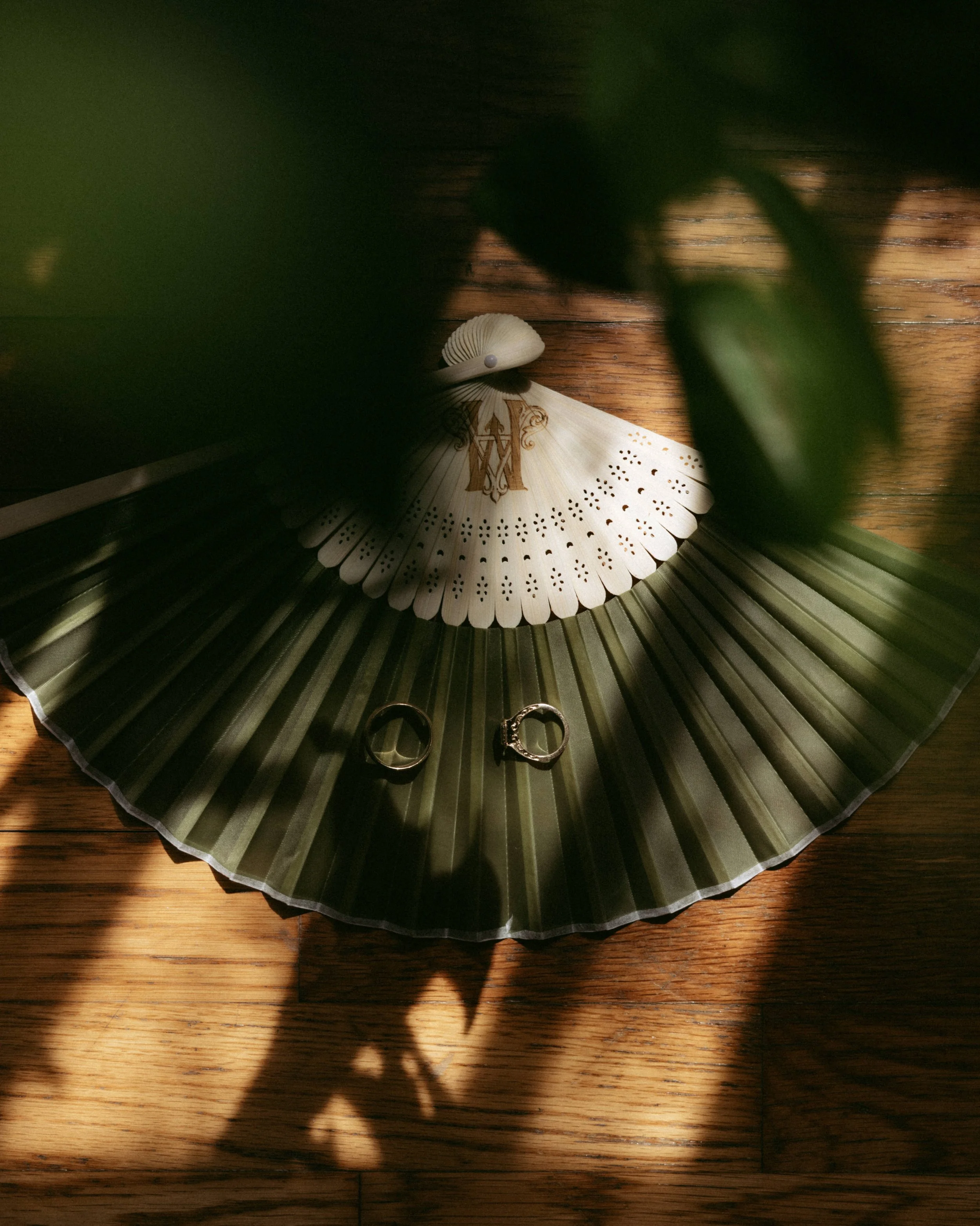 A wooden surface with a decorative paper fan, a silver ring, and a gold ring on top. A blurred plant is in the foreground, casting a shadow on the surface.