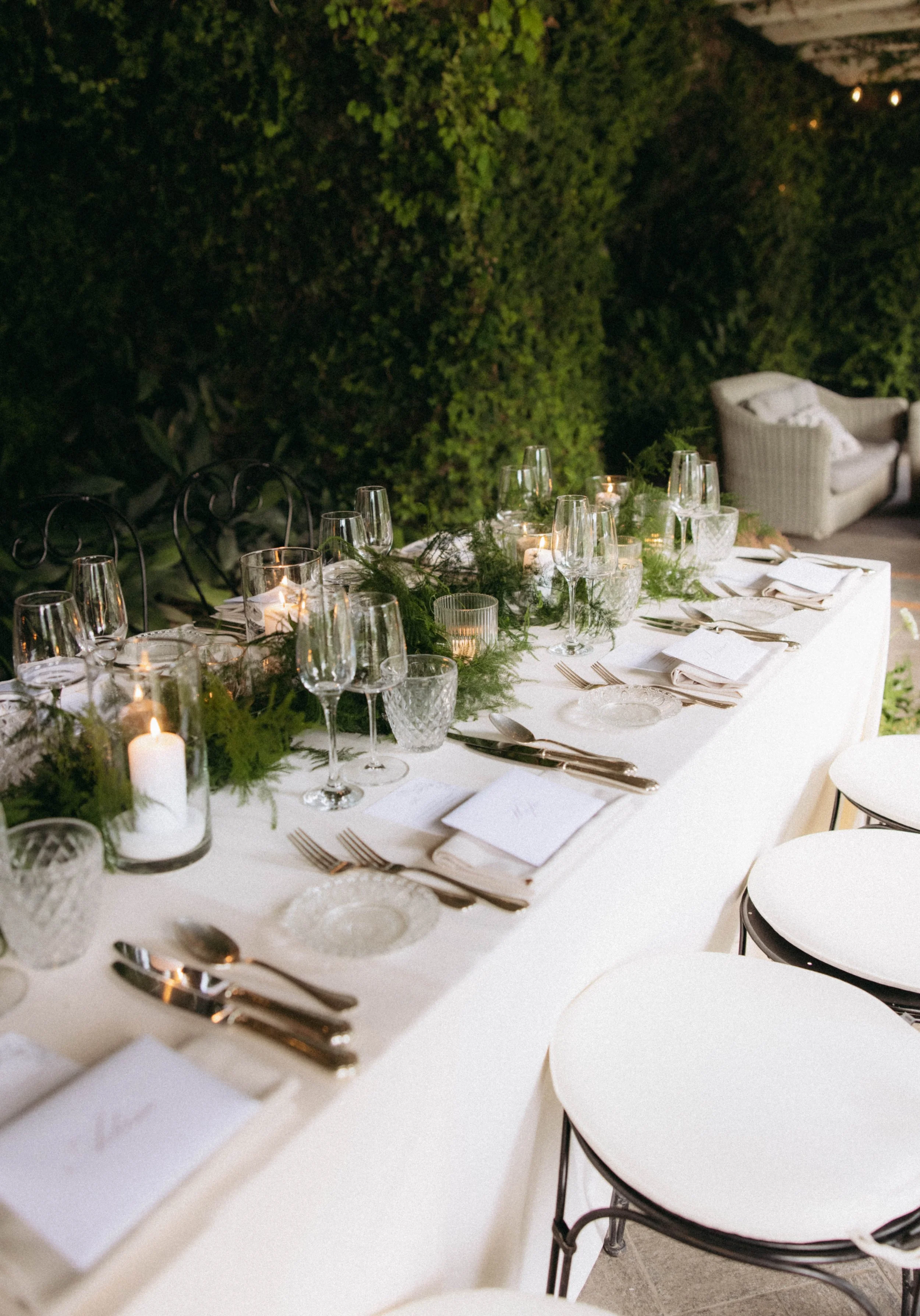 A dinner table set outdoors for a formal event, with white tablecloth, multiple wine glasses, silverware, white napkins, candles, and greenery decorations, with a white couch and lush green hedge in the background.