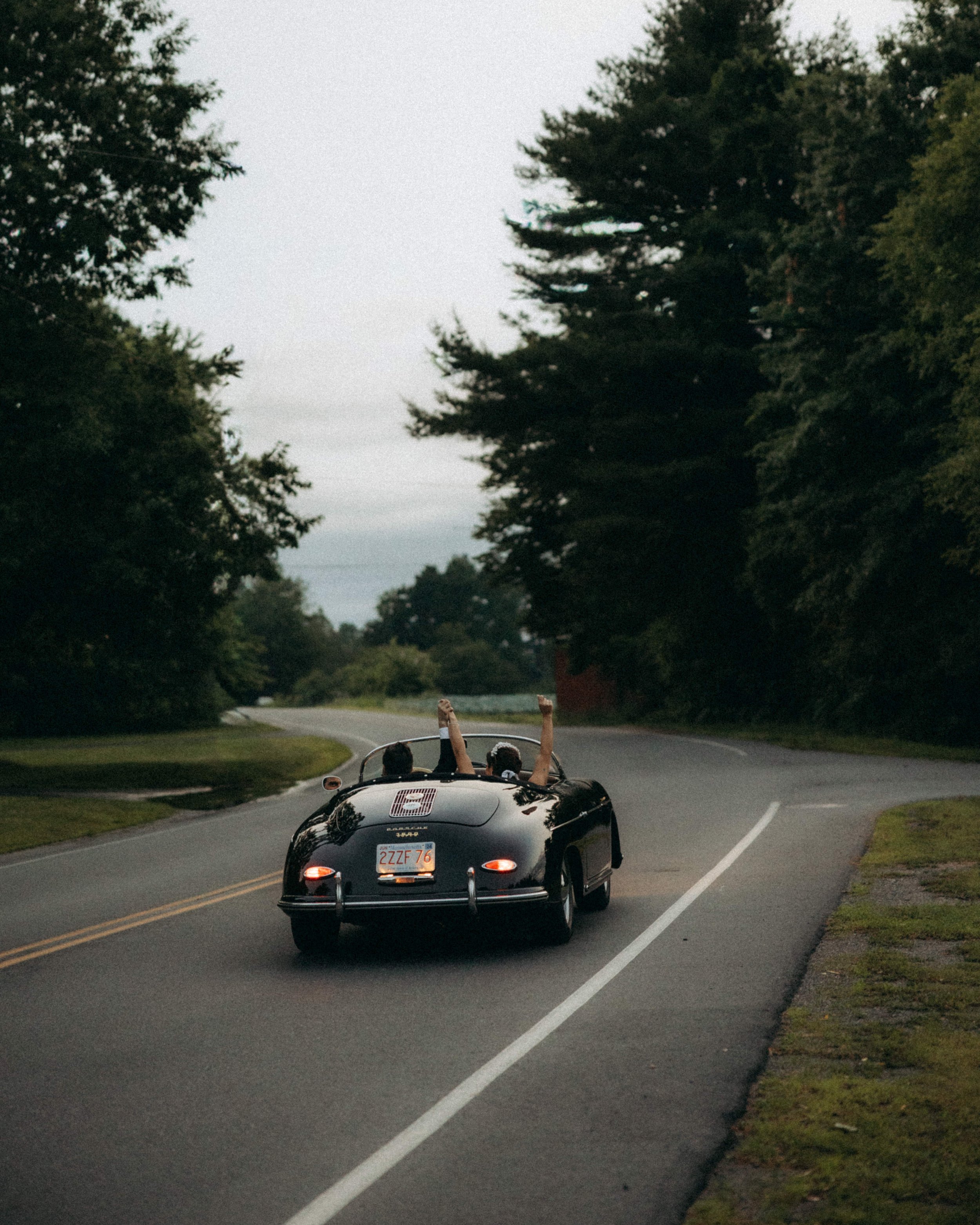 A black convertible sports car with a Massachusetts license plate driving down a winding road surrounded by greenery and trees, with two children in the passenger and driver seats raising their arms.