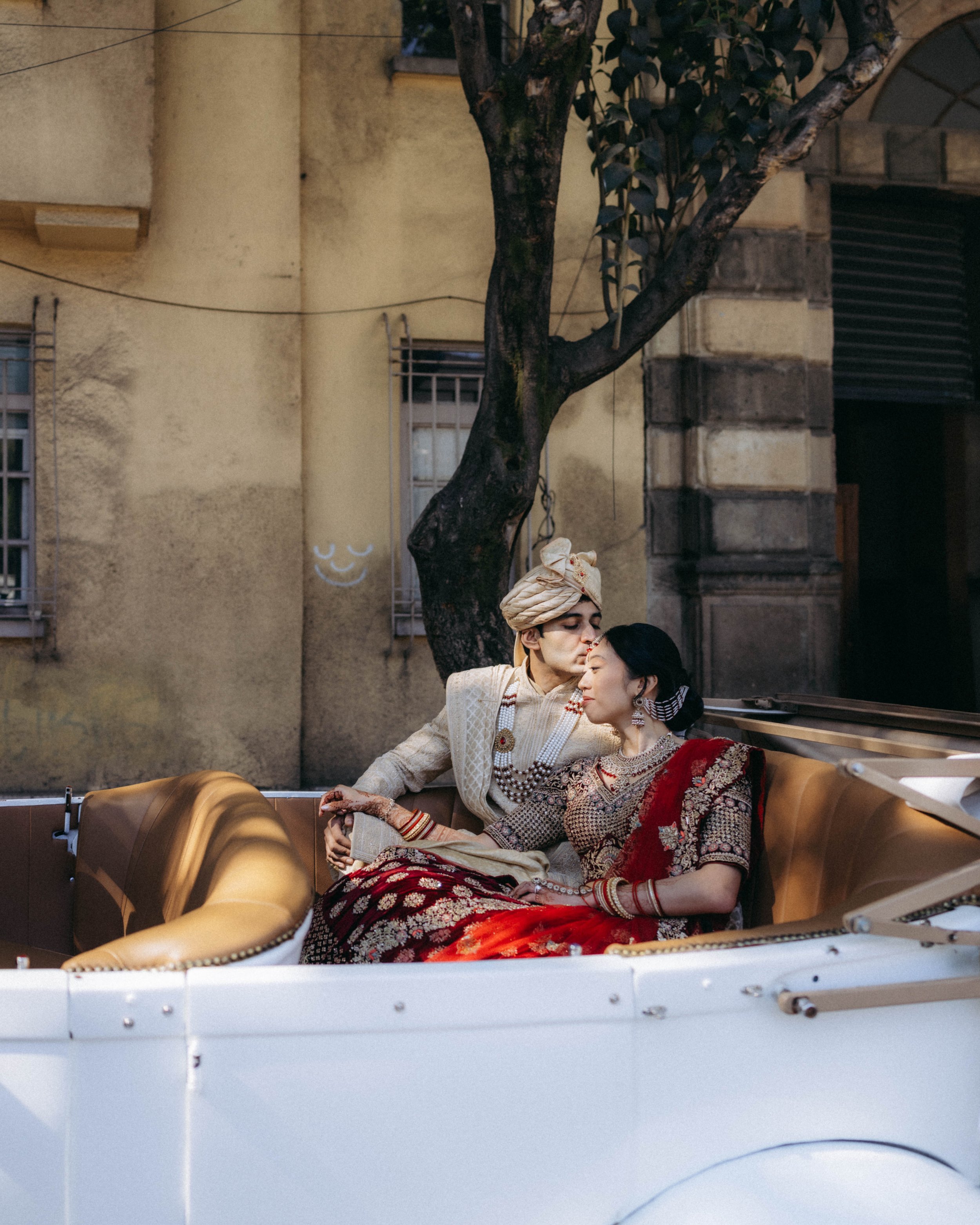 A couple dressed in traditional Indian attire sitting closely together on a vintage car, sharing an intimate moment, with an old yellow wall and a tree in the background.