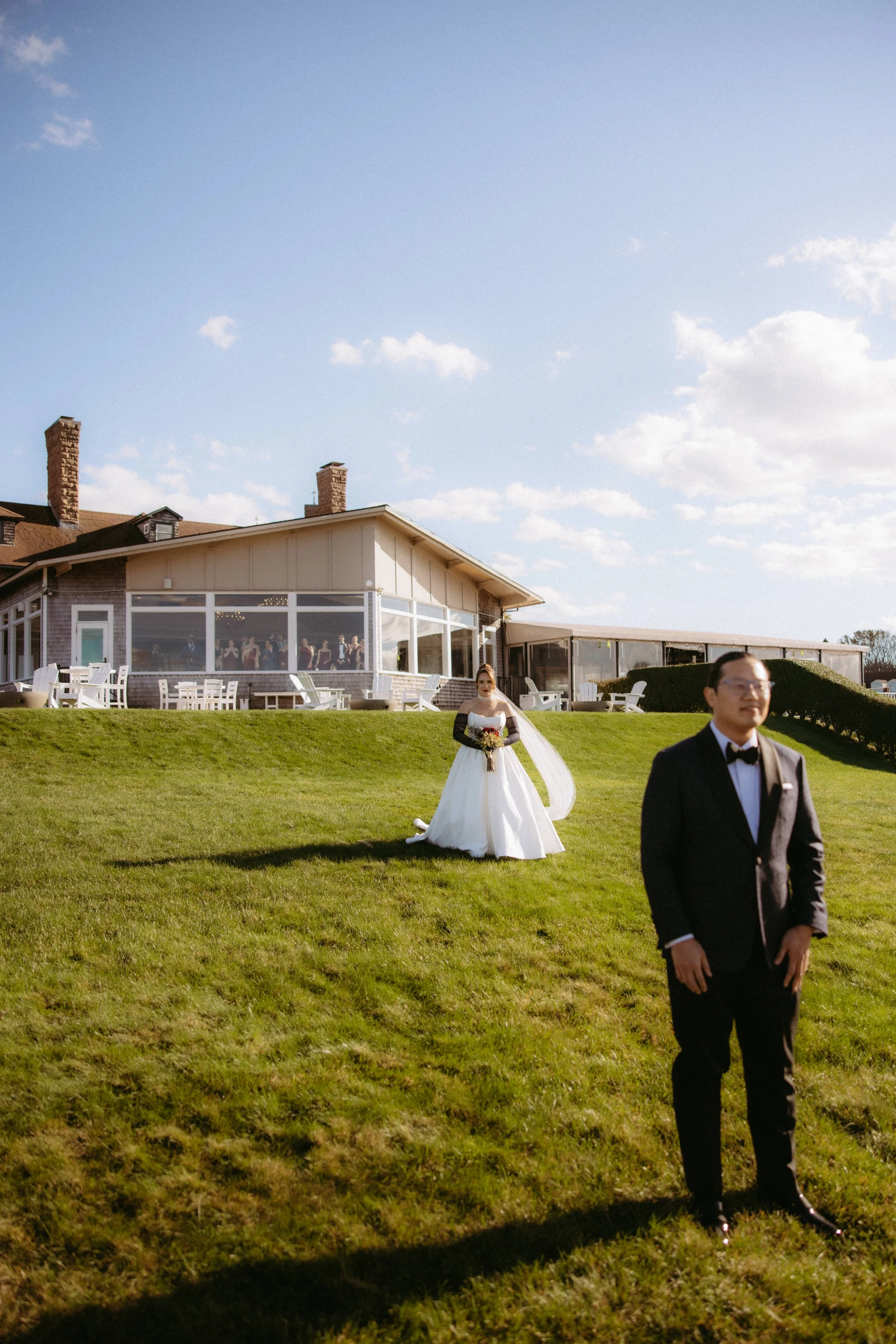A bride in a white wedding dress holding a bouquet and a groom in a tuxedo walking on a grassy lawn outside a building with a glass enclosure.