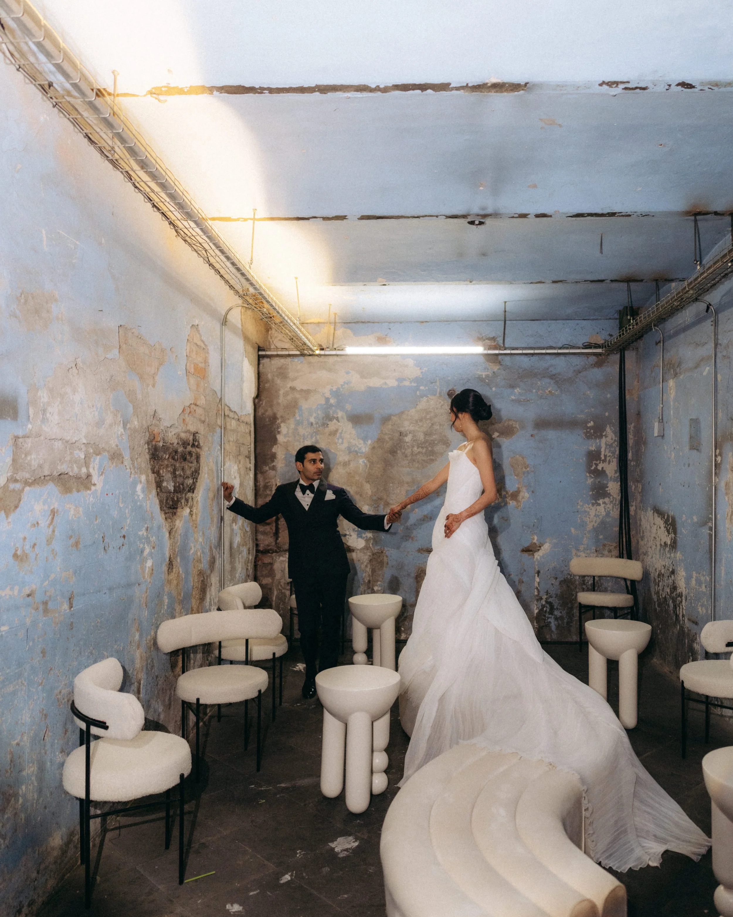 A woman in a white wedding dress and a man in a tuxedo holding hands in an industrial space with peeling paint walls and chairs, a curved marble or stone structure, and exposed lighting fixtures.