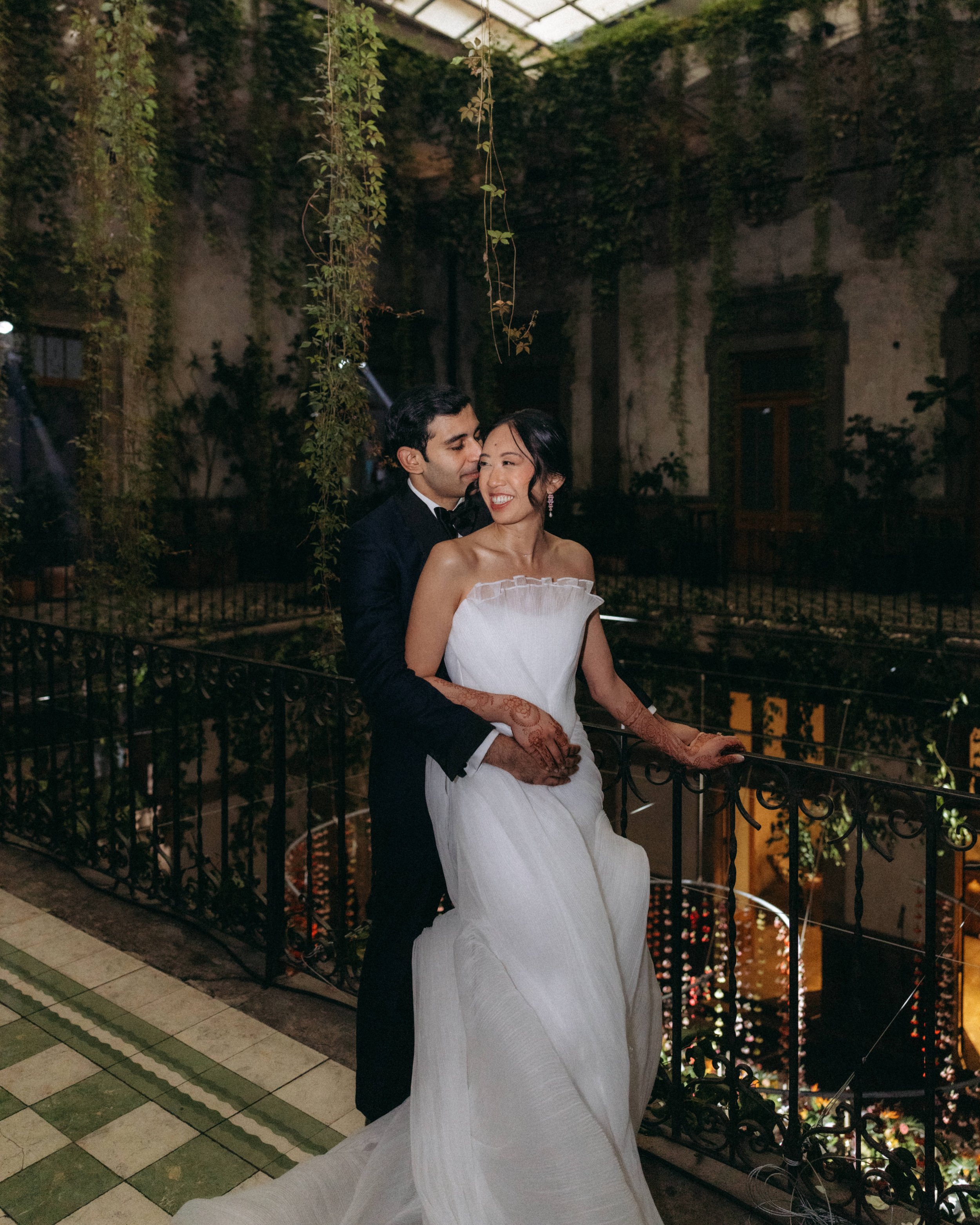 A couple dressed in formal attire, the man in a tuxedo and the woman in a white dress, embracing and smiling at each other on an outdoor balcony at night, decorated with hanging vines and warm string lights.