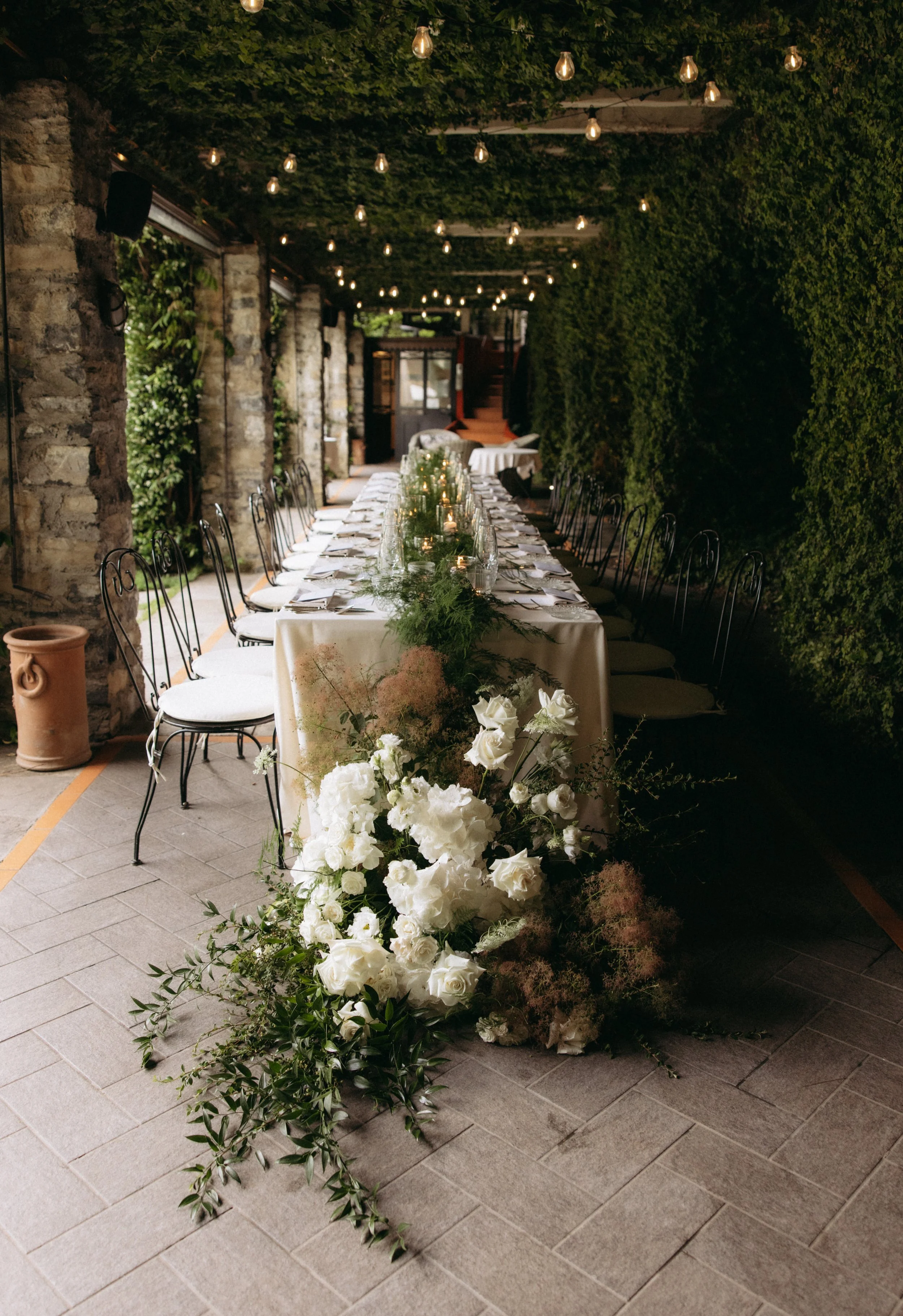 Long dining table decorated with white flowers and greenery, set for an event or party, in a covered outdoor space with hanging lights, surrounded by stone walls and lush greenery.