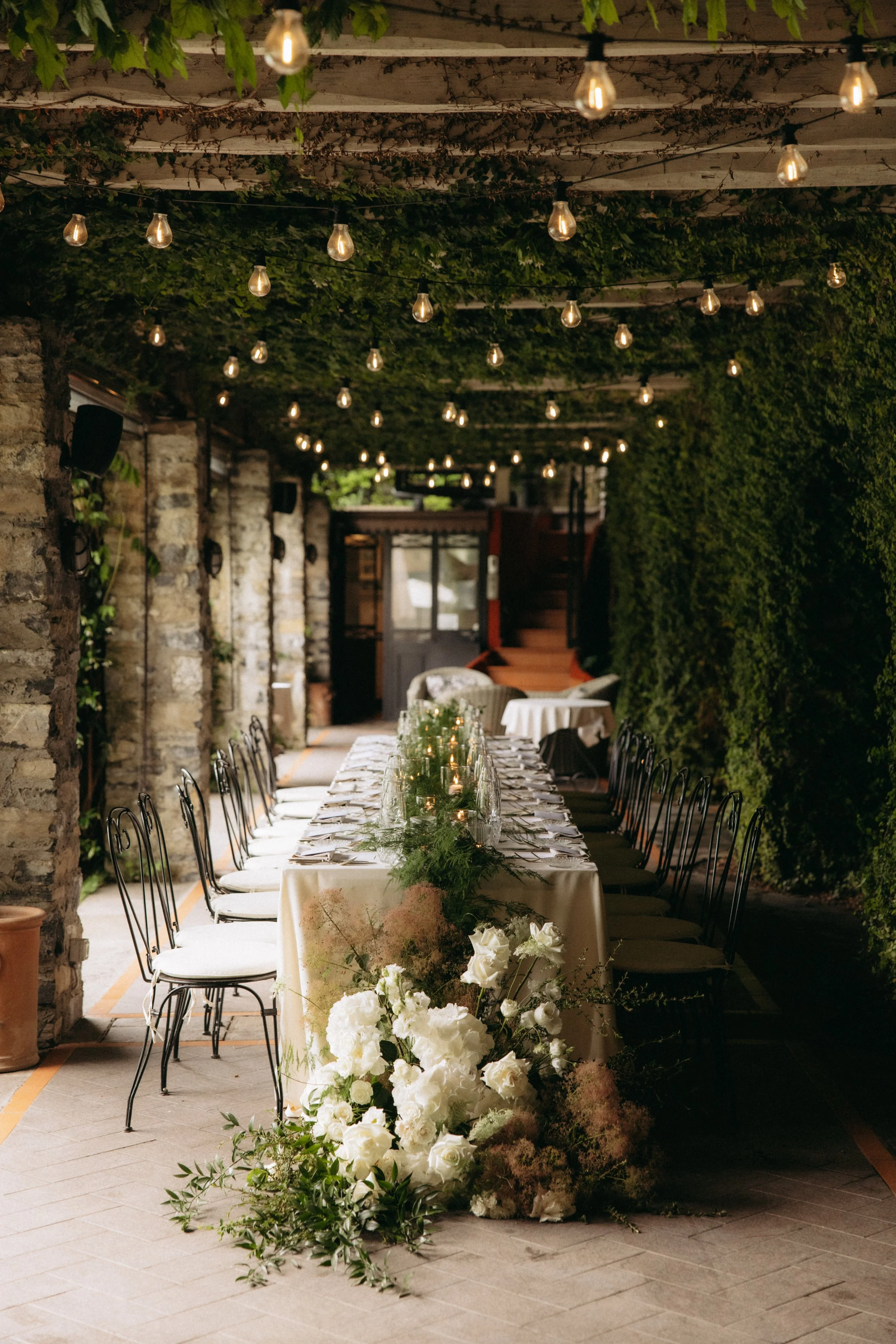 Decorated outdoor dining area with a long table, white tablecloth, floral arrangements, and overhead string lights, set under a green leafy canopy at dusk.